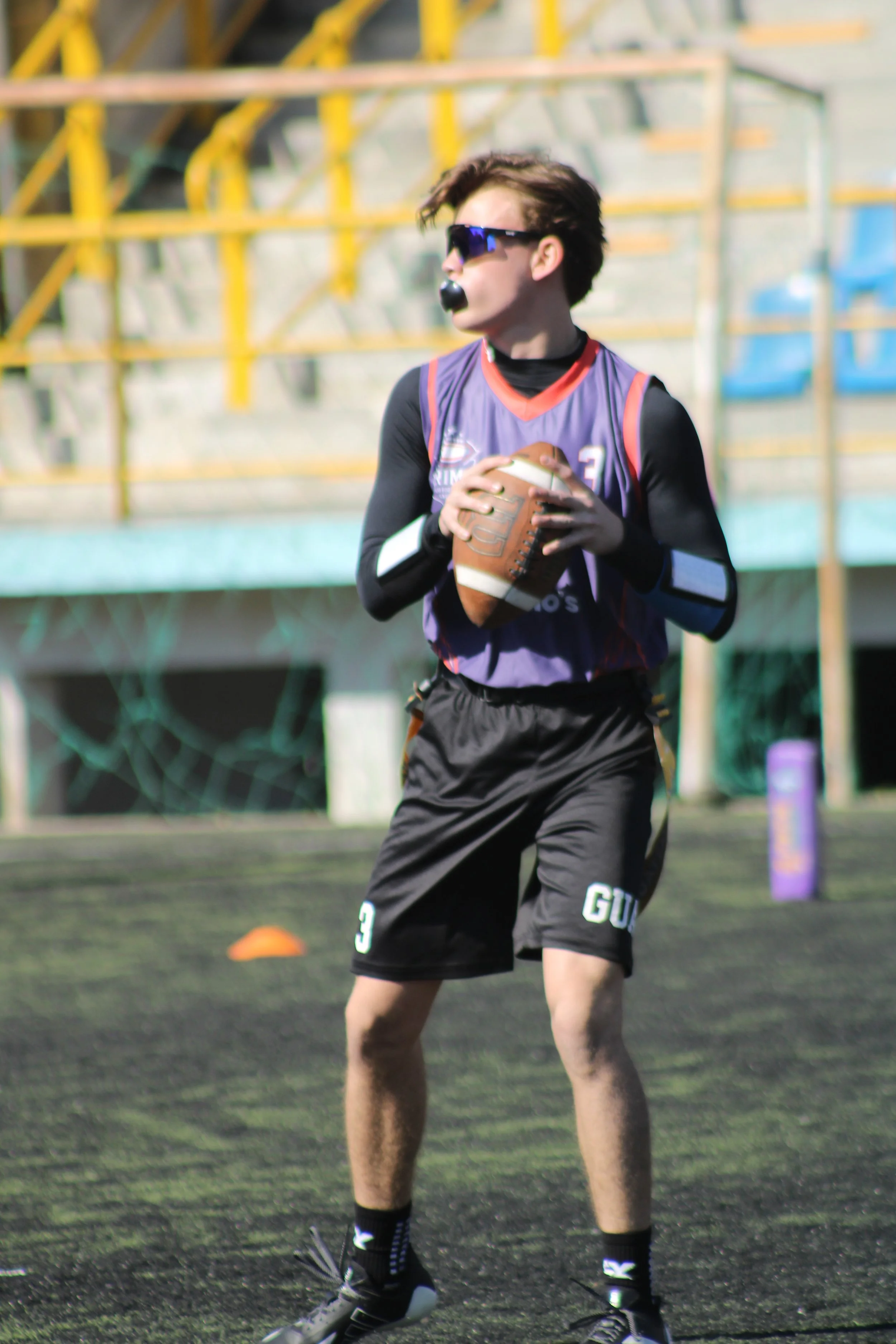 A young person with brown hair, wearing sunglasses, a purple sports jersey, black shorts with the number 9, and black gloves, holding a football on a field.