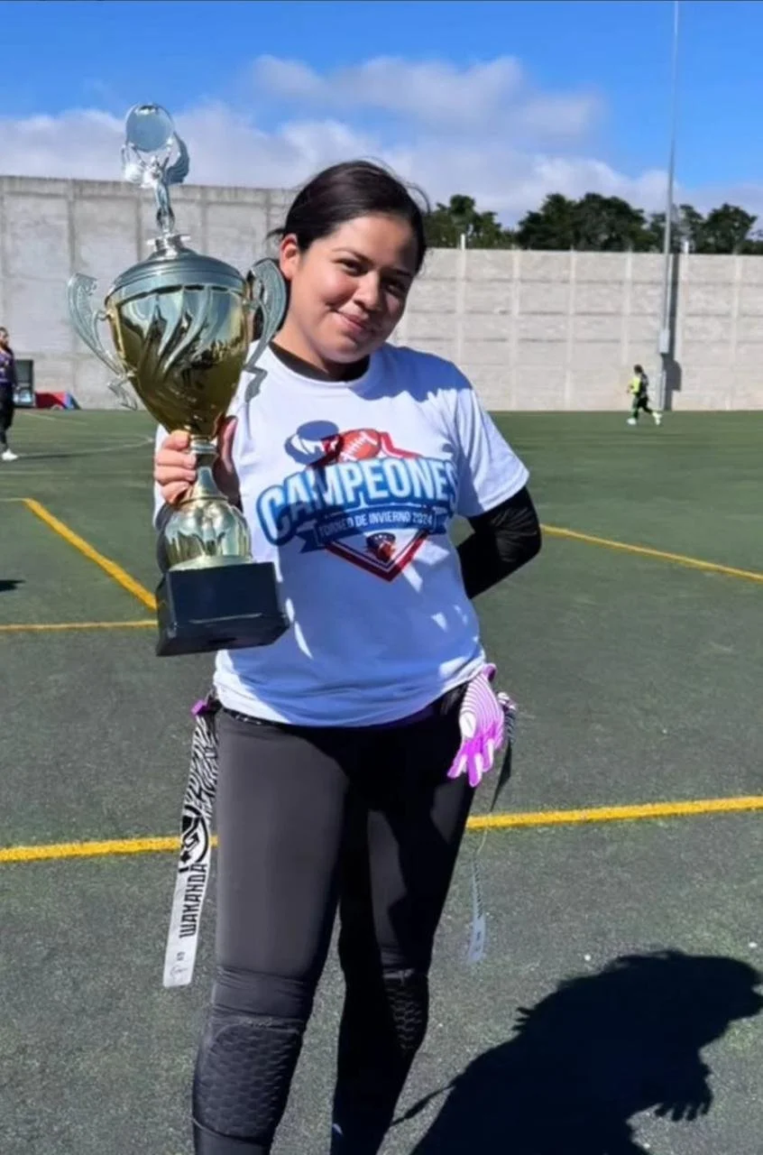 A young woman standing on a sports field holding a large trophy, wearing a white t-shirt with a 'Campeones' logo, and smiling at the camera.