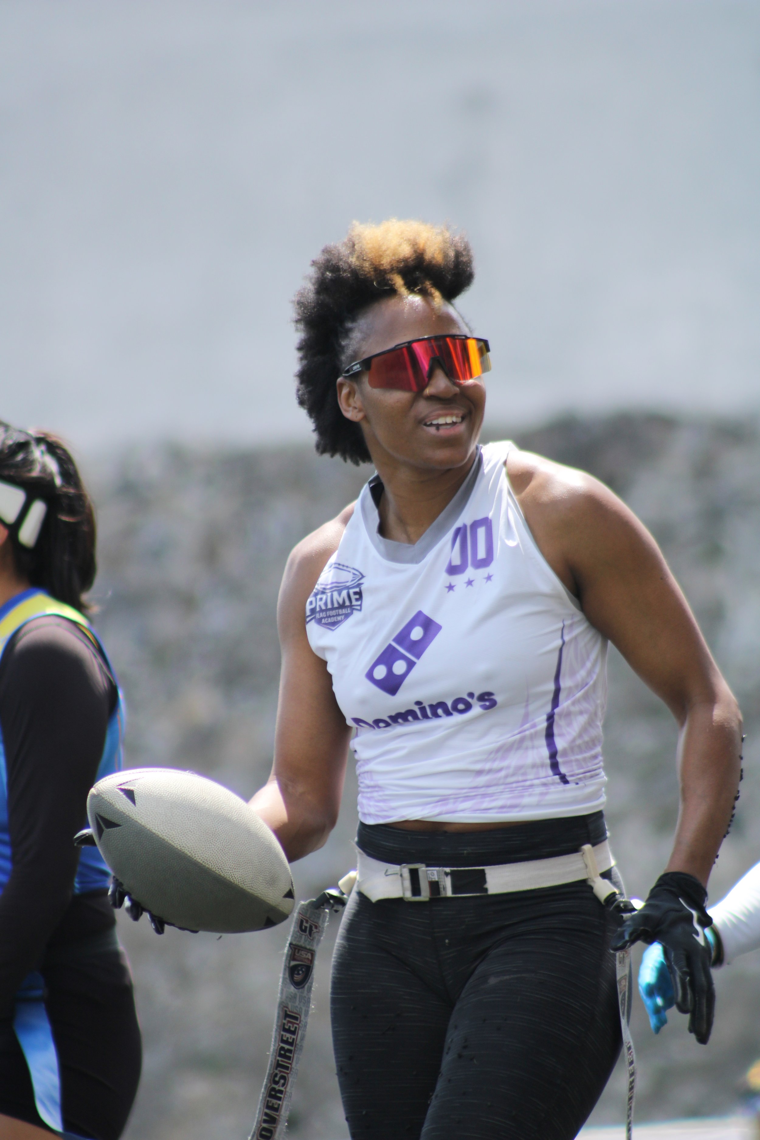 Female athlete with curly hair, wearing sunglasses and sportswear, holding a football, outdoors during a sporting event.