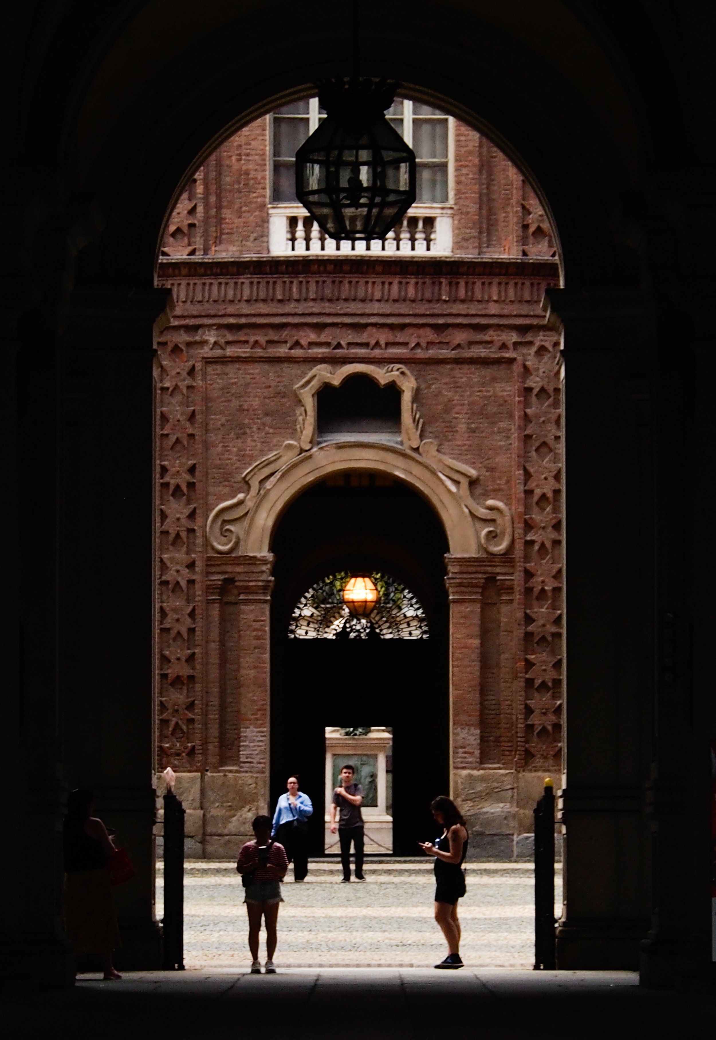 People standing and walking through a large arched entrance with ornate brick walls and decorative features, looking into a courtyard with a lamp and building details in the background.
