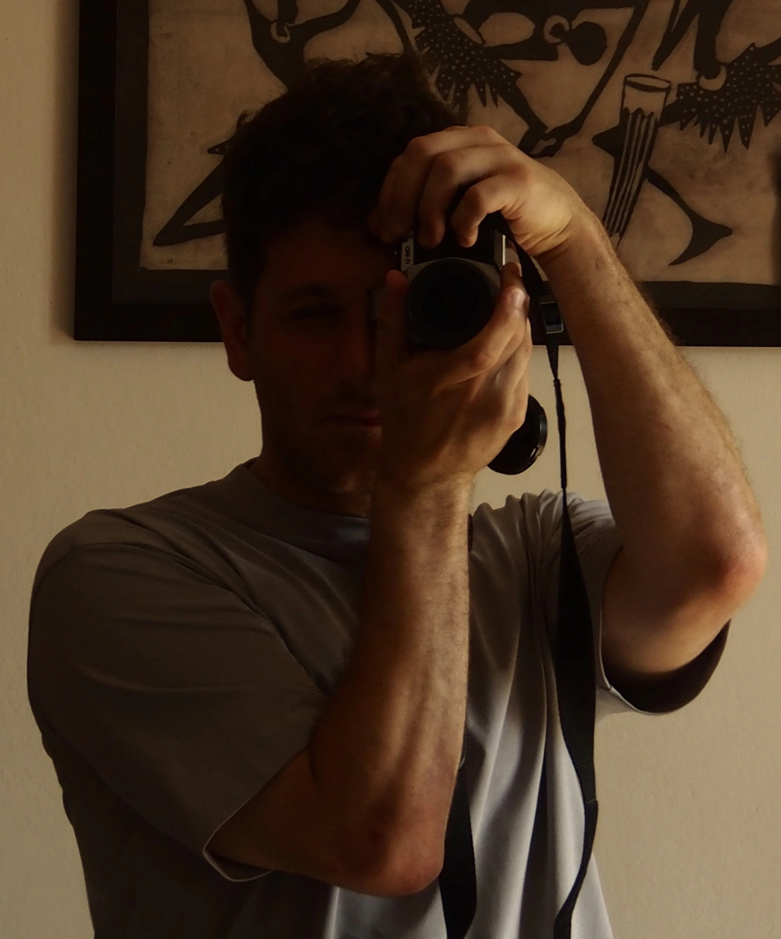 A man taking a photo of himself in a mirror with a camera, standing in front of a framed painting or illustration of fish.