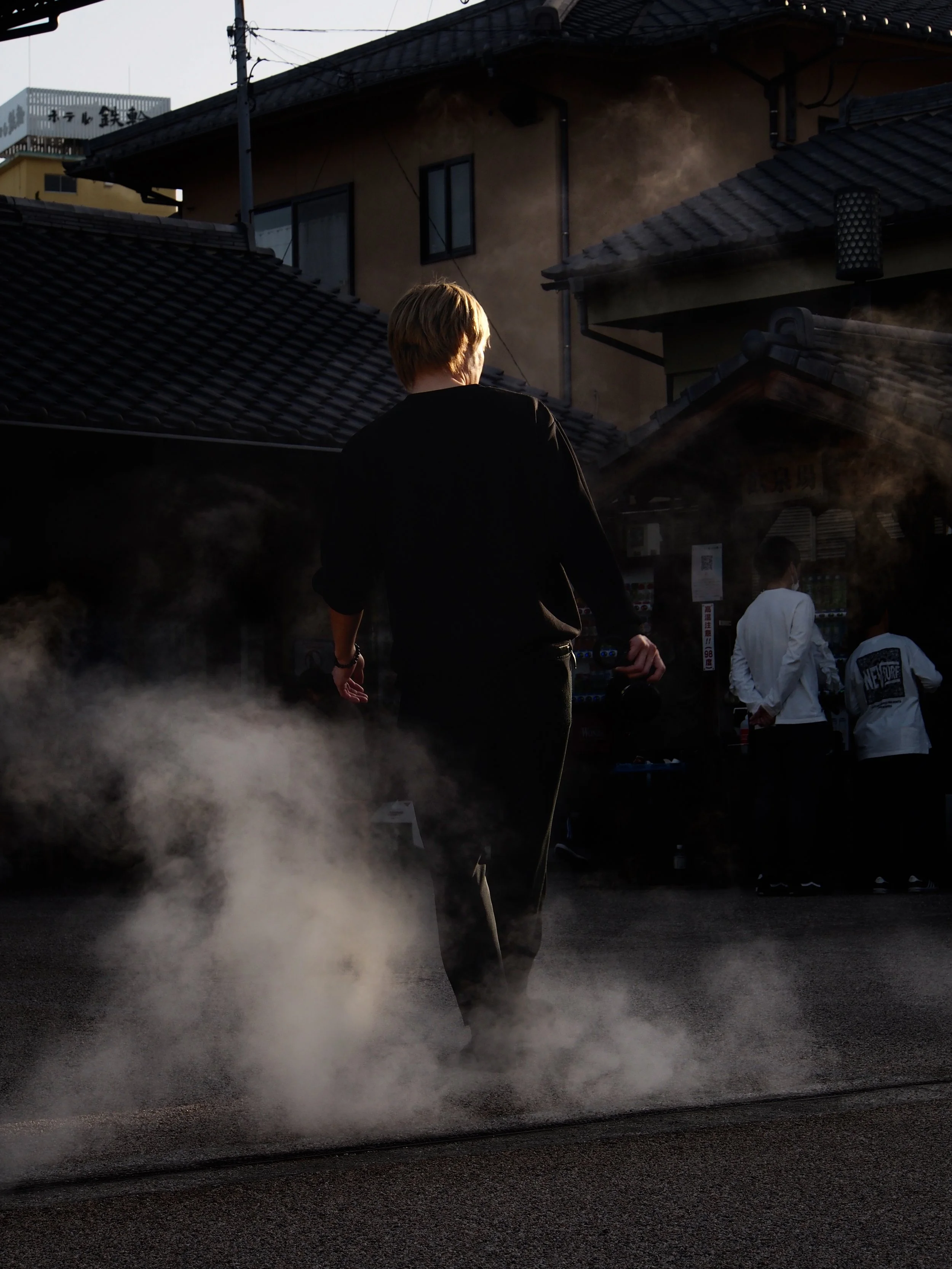 A person walking away from the camera on a foggy street at sunset, with traditional Japanese-style buildings and other people in the background.