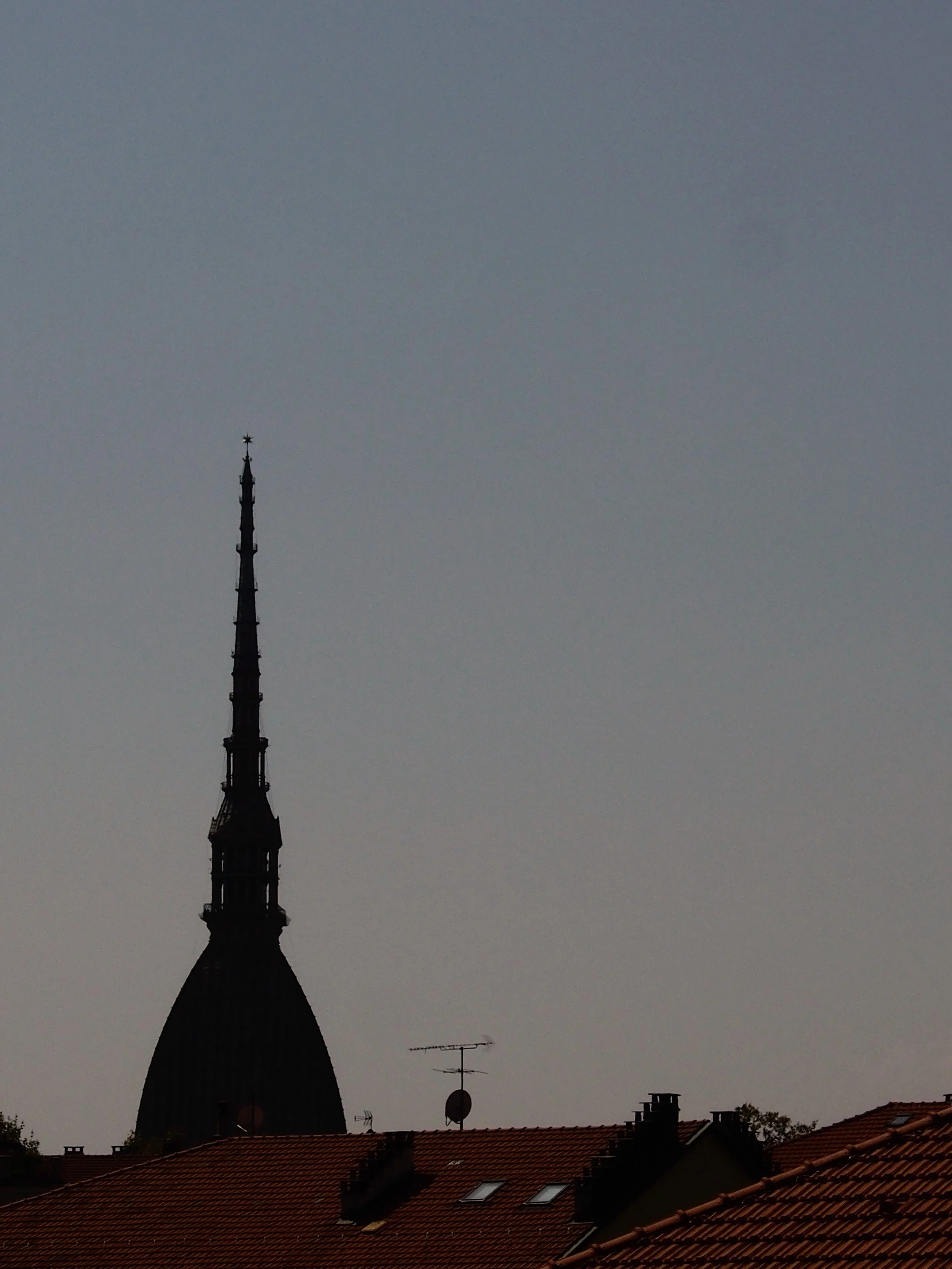 Silhouette of a tall church steeple with a star at the top, viewed over rooftops with red tiles, against a clear sky at dusk or dawn.
