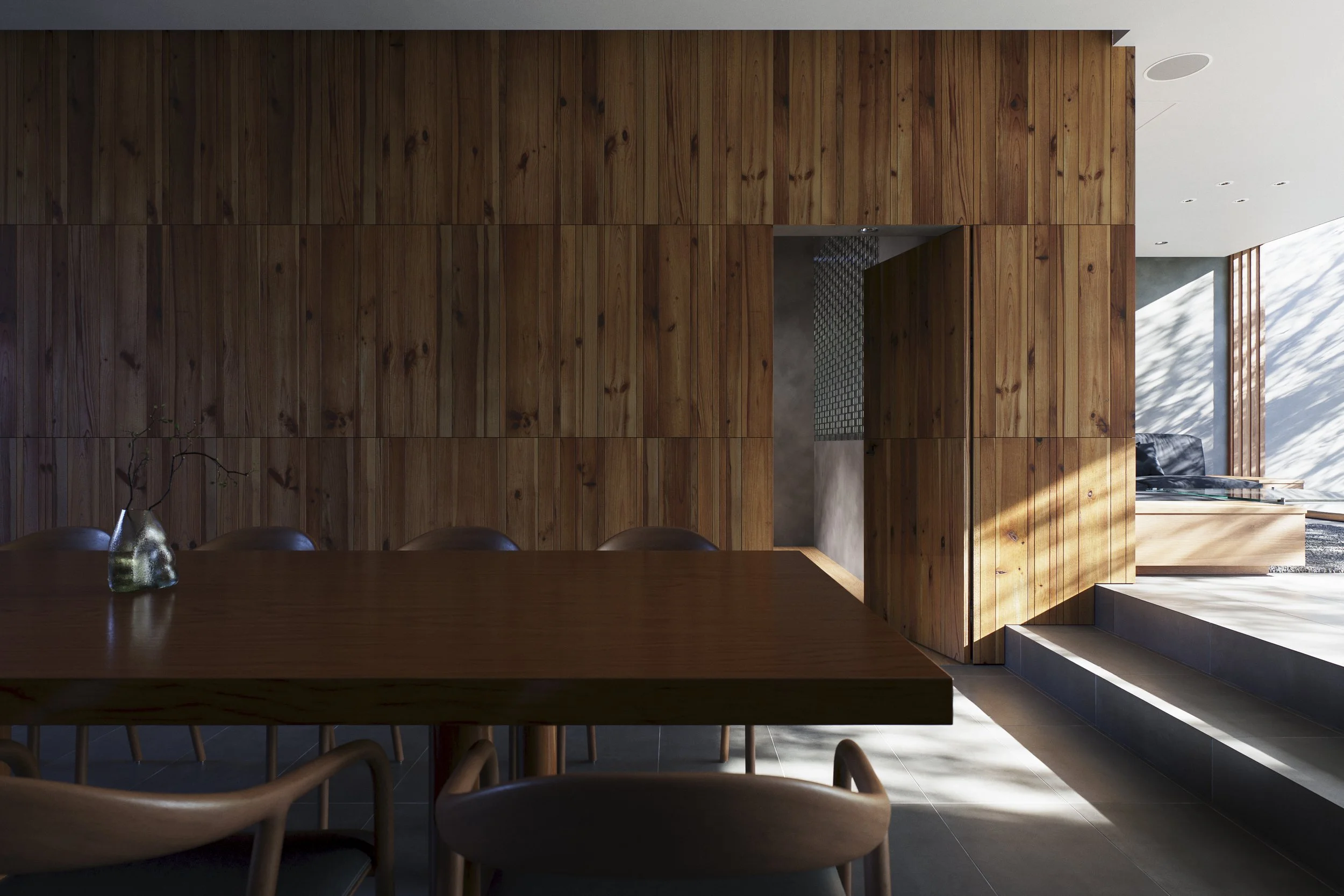 Interior of a modern home with a large wooden wall, a dining table with chairs, and a view into a living area with a black sofa and large windows letting in natural light.