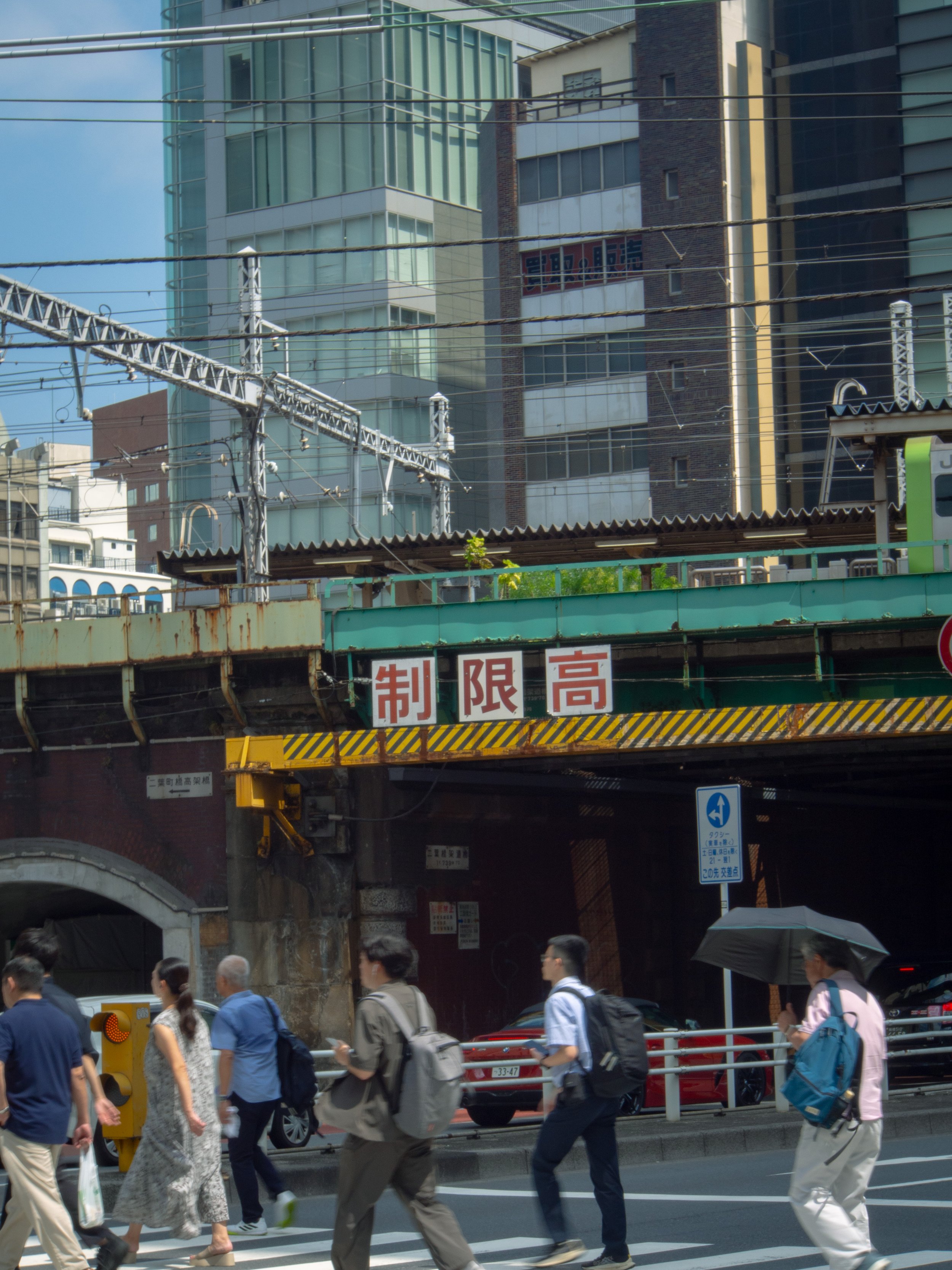 People crossing the street beneath an overpass with Japanese sign. Urban cityscape with tall buildings and train tracks overhead.