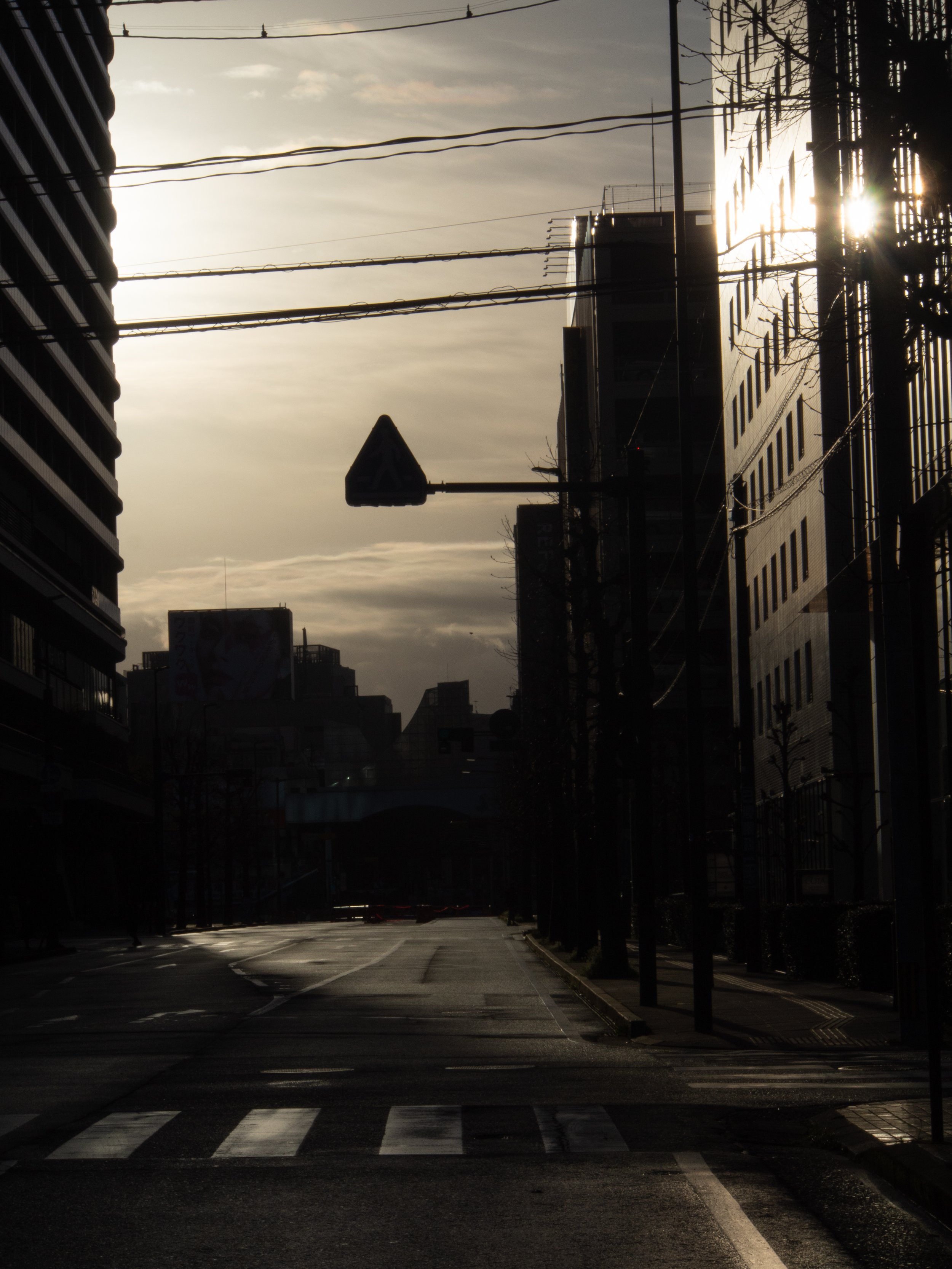 A city street at sunset with tall buildings, power lines, a traffic signal, and a pedestrian crosswalk.