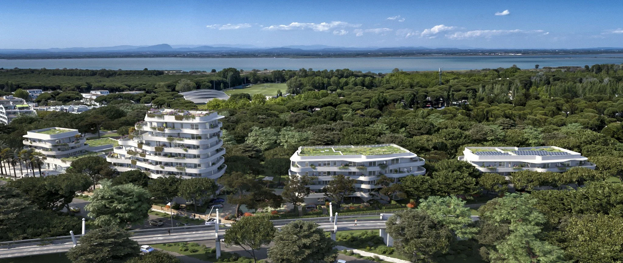 Modern white residential buildings with curved balconies surrounded by lush green trees, set against a backdrop of a large body of water and distant mountains under a partly cloudy sky.