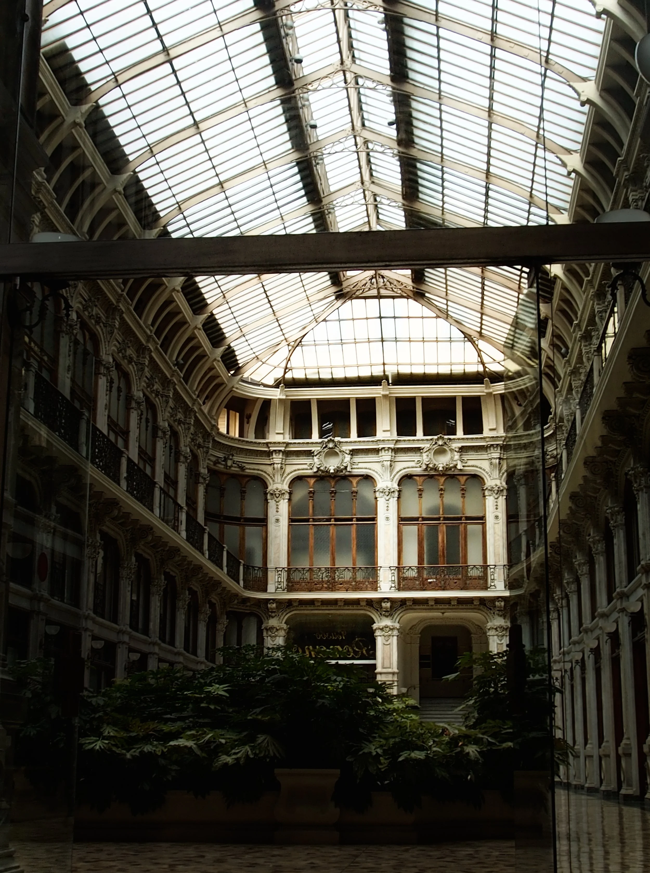 Interior of a historic shopping arcade or gallery with a glass ceiling, ornate architectural details, and plants in the center.