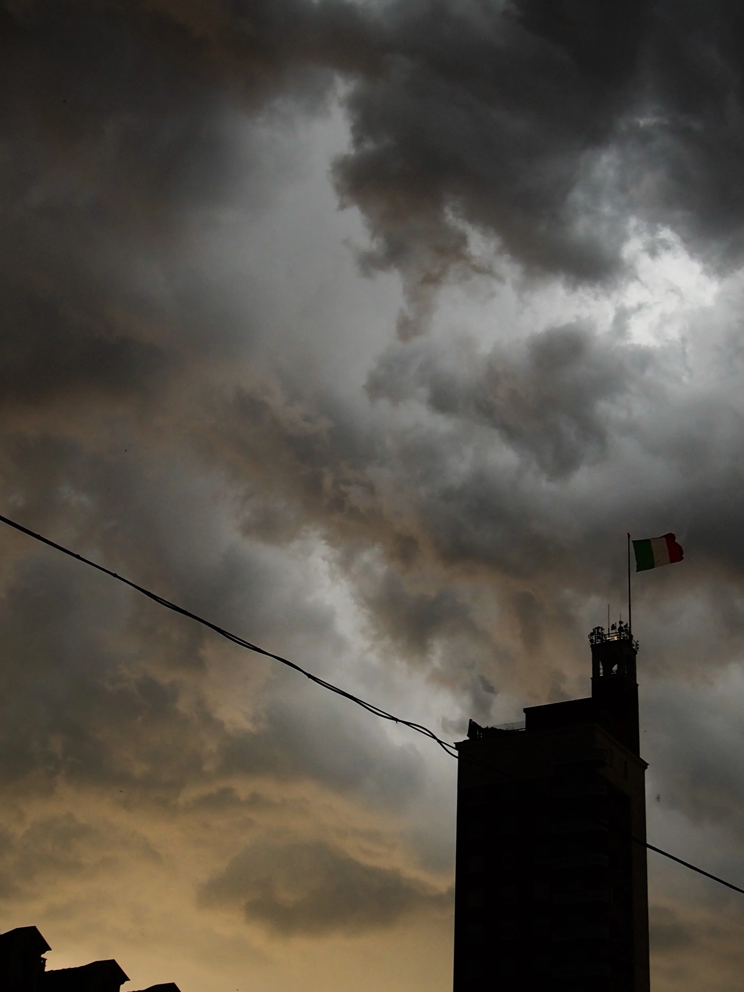 Dark, cloudy sky over a city with a tall building flying an Italian flag.