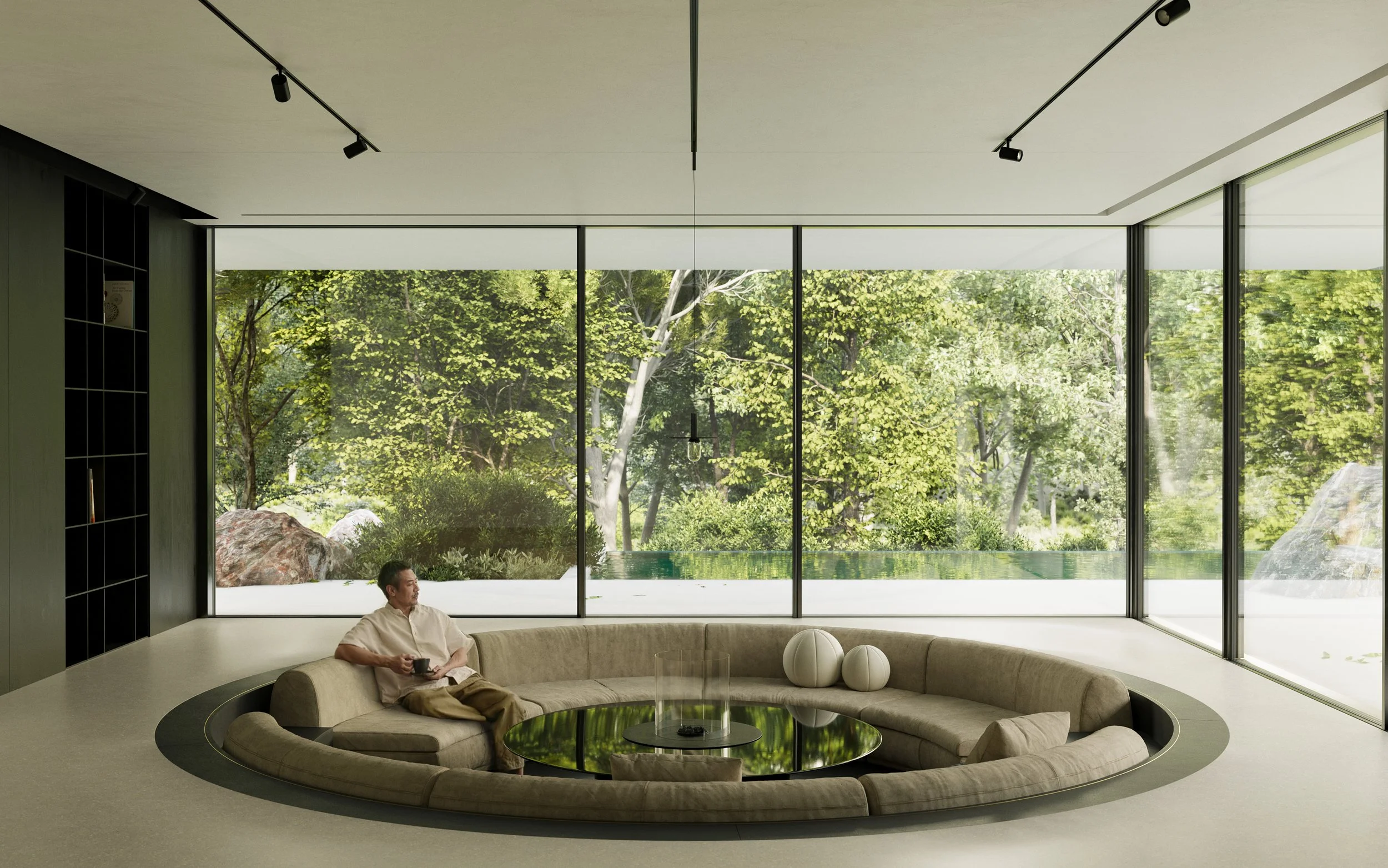 A man sitting on a circular beige sofa inside a modern room with large floor-to-ceiling glass windows showing a lush green outdoor landscape.
