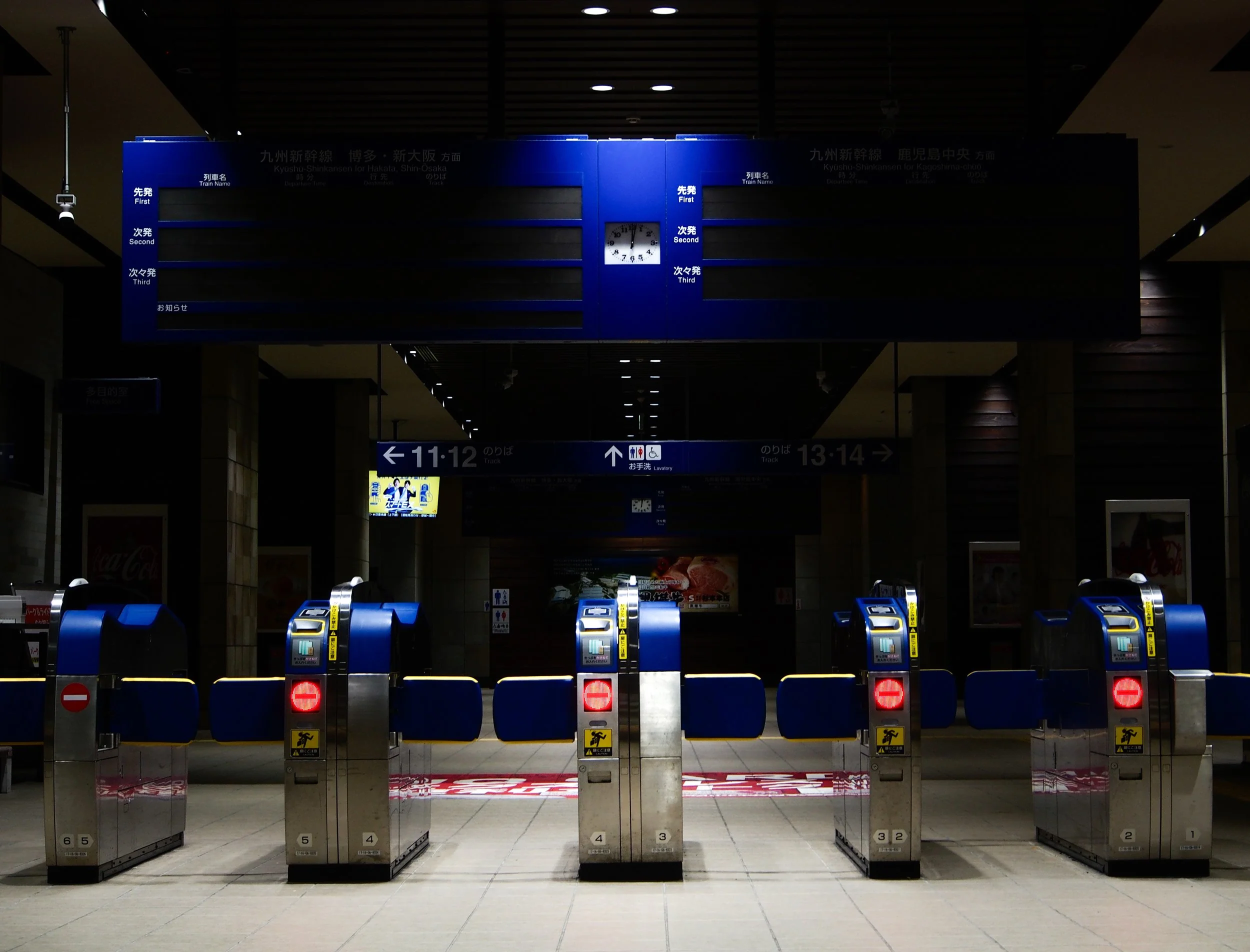 Empty train station entrance with ticket gates, blue barriers, and a digital departure board overhead showing train information.