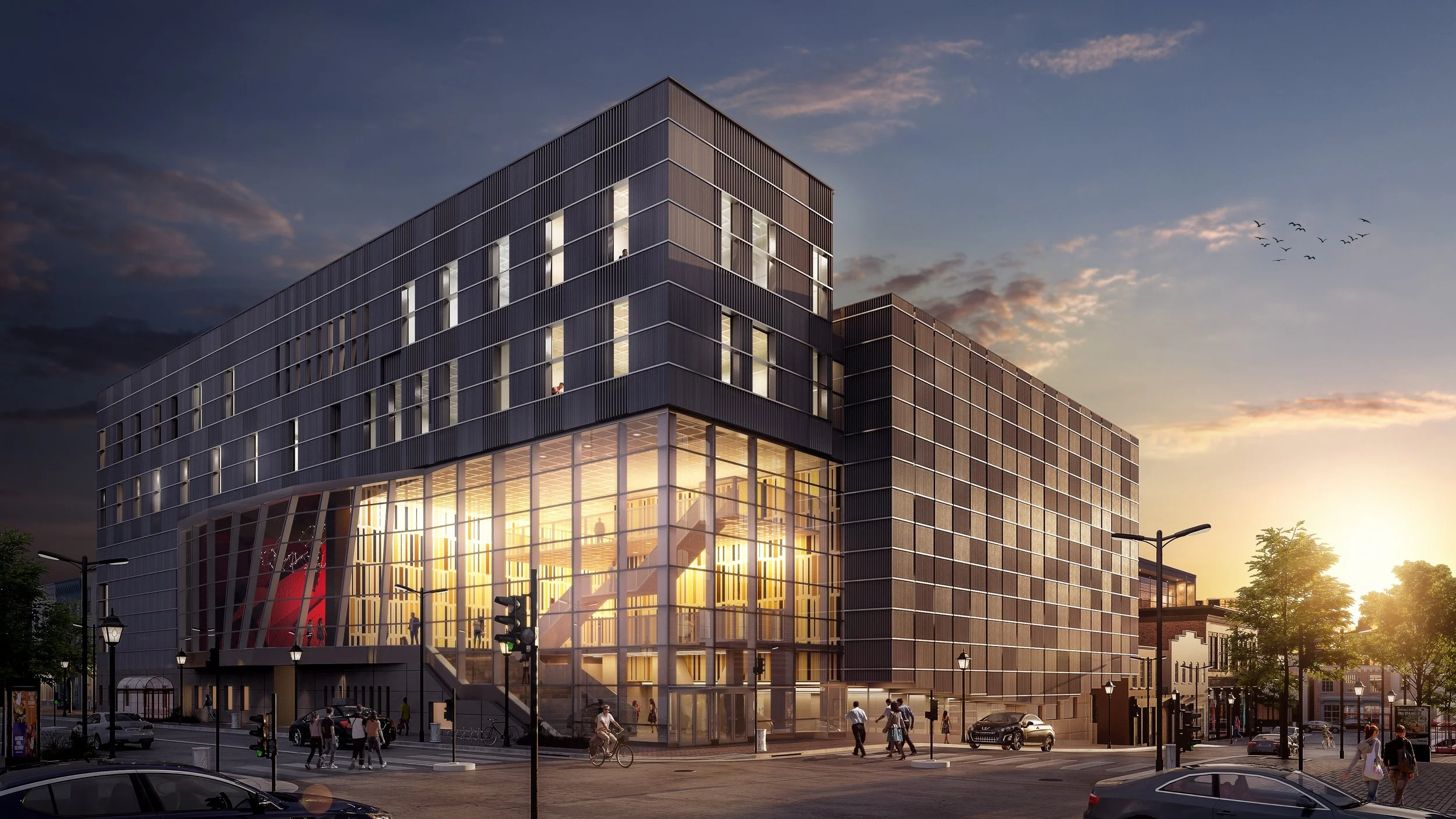 Modern multi-story building with large glass windows and exterior lighting during sunset, with cars and pedestrians on the street.