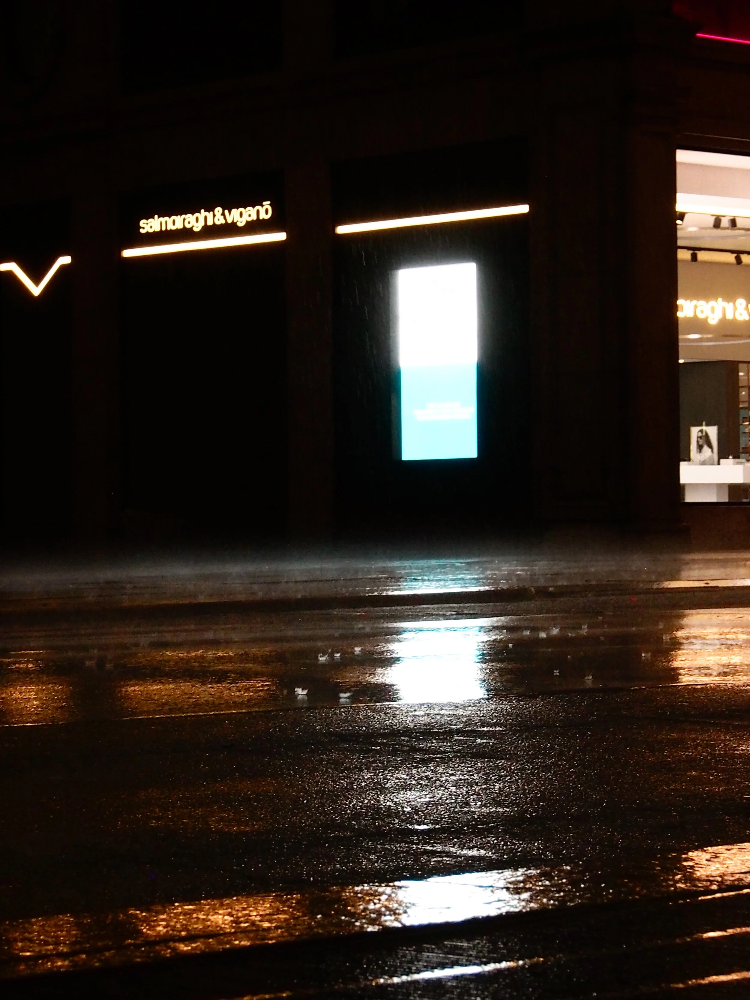 Nighttime scene showing wet street reflecting lights from storefronts, including a Dior store and a sign for Salvatore Ferragamo & Viganò, with a digital screen on the sidewalk.