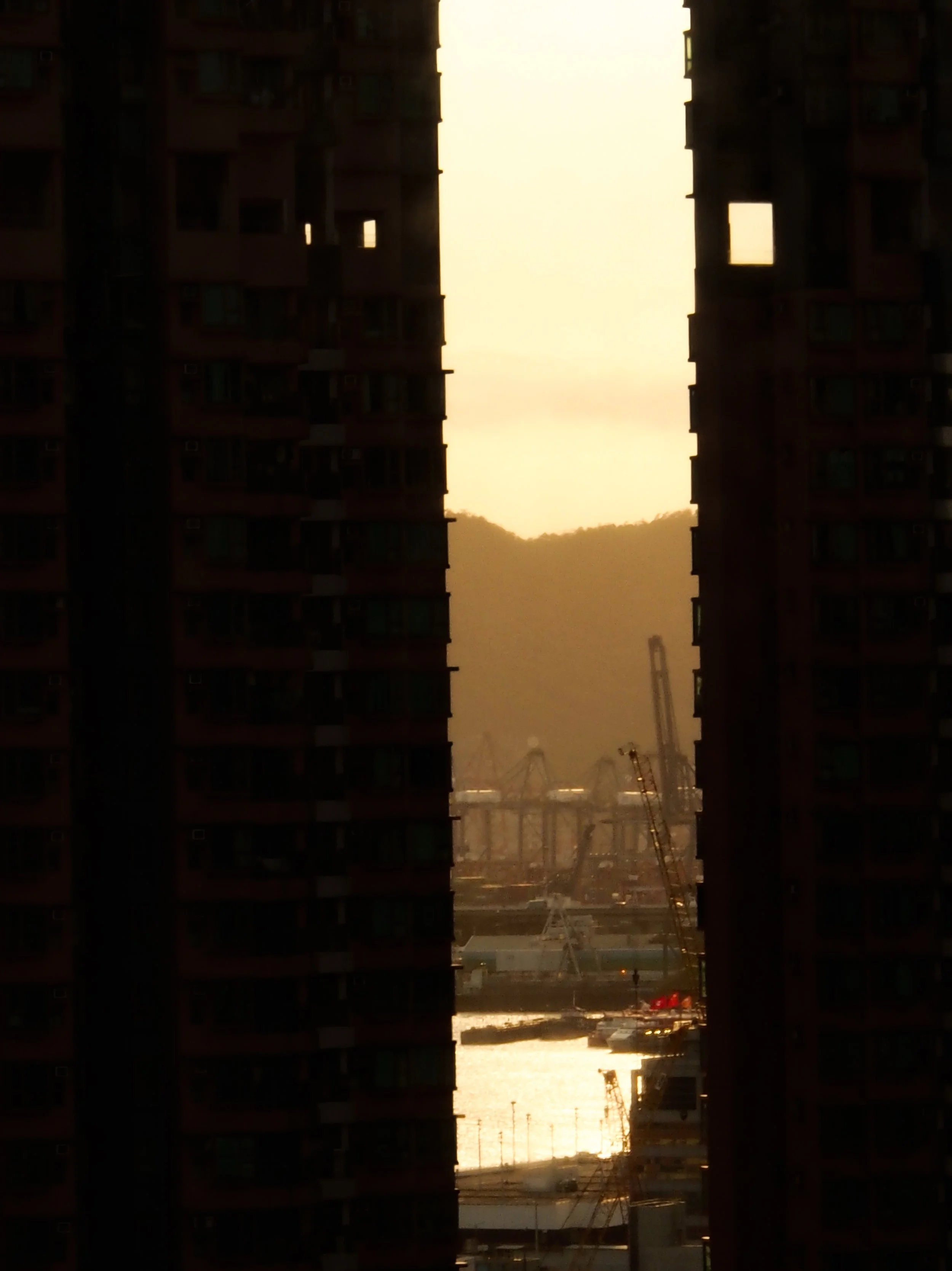 A cityscape viewed through a narrow gap between two tall dark buildings, with a crane and industrial structures near the water, and a mountain in the background during sunset.