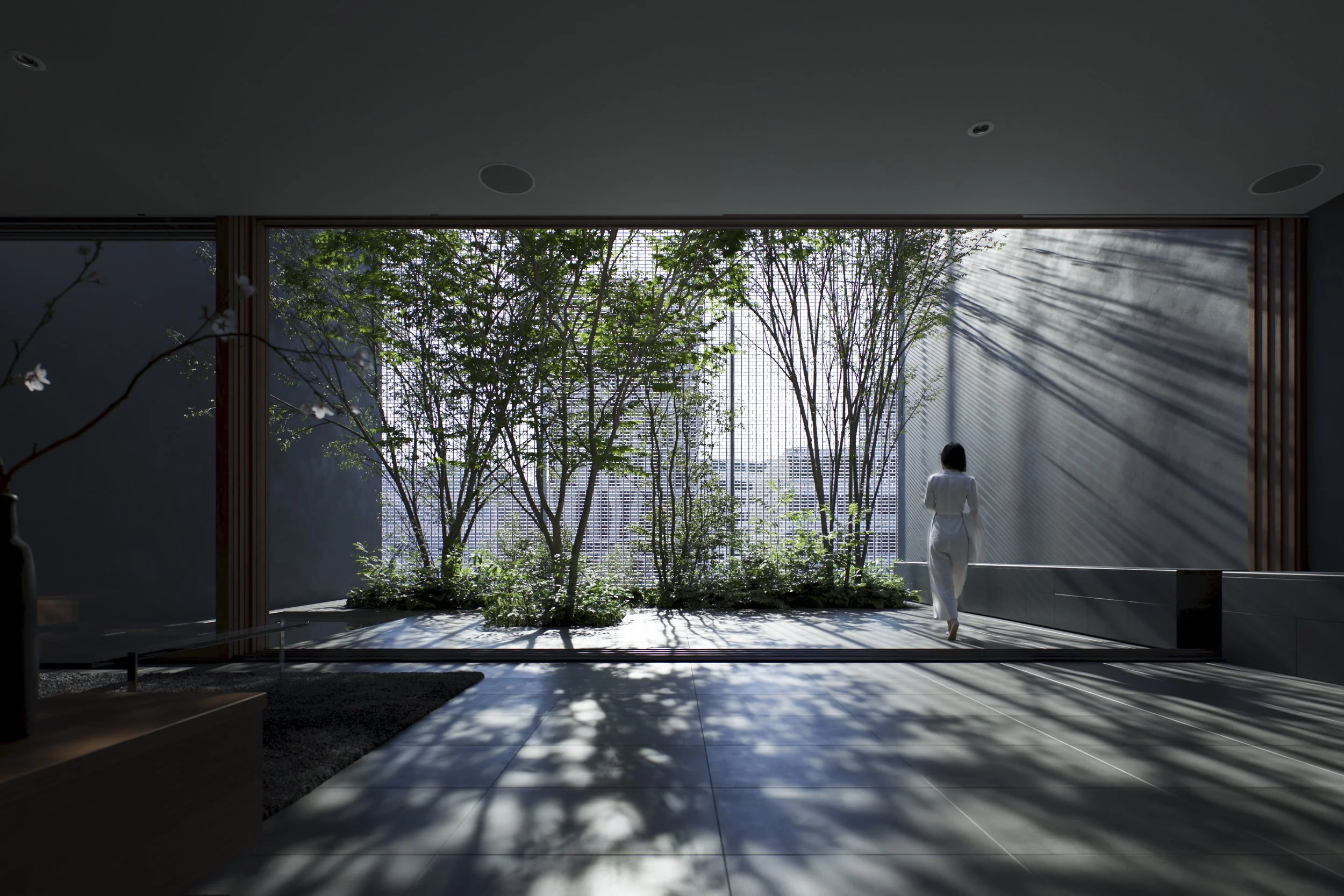 Interior view of a modern building with large windows, featuring a person in white walking by a lush indoor garden with tall trees, and sunlight casting shadows on the tiled floor.