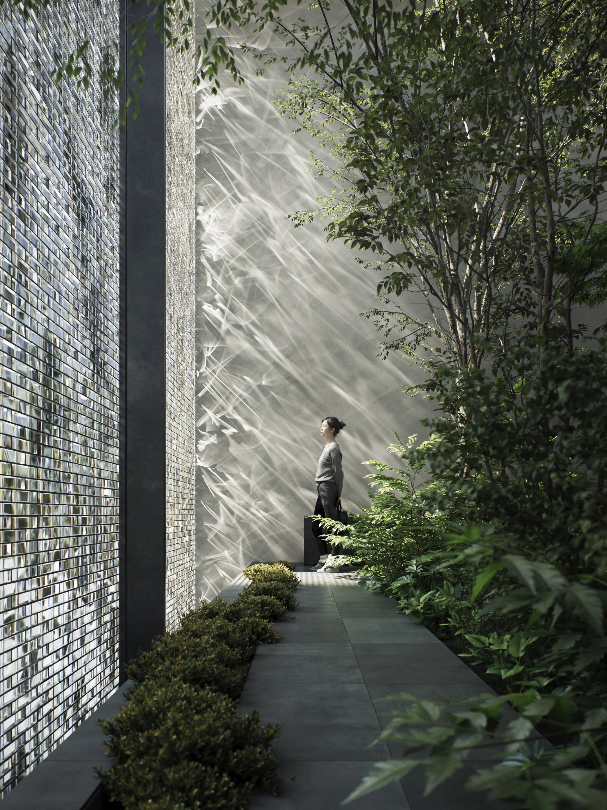 A woman standing on a narrow outdoor pathway lined with plants on both sides and a modern wall with a mosaic tile pattern on the left. She holds a black bag and looks up toward the sunlight filtering through the trees.