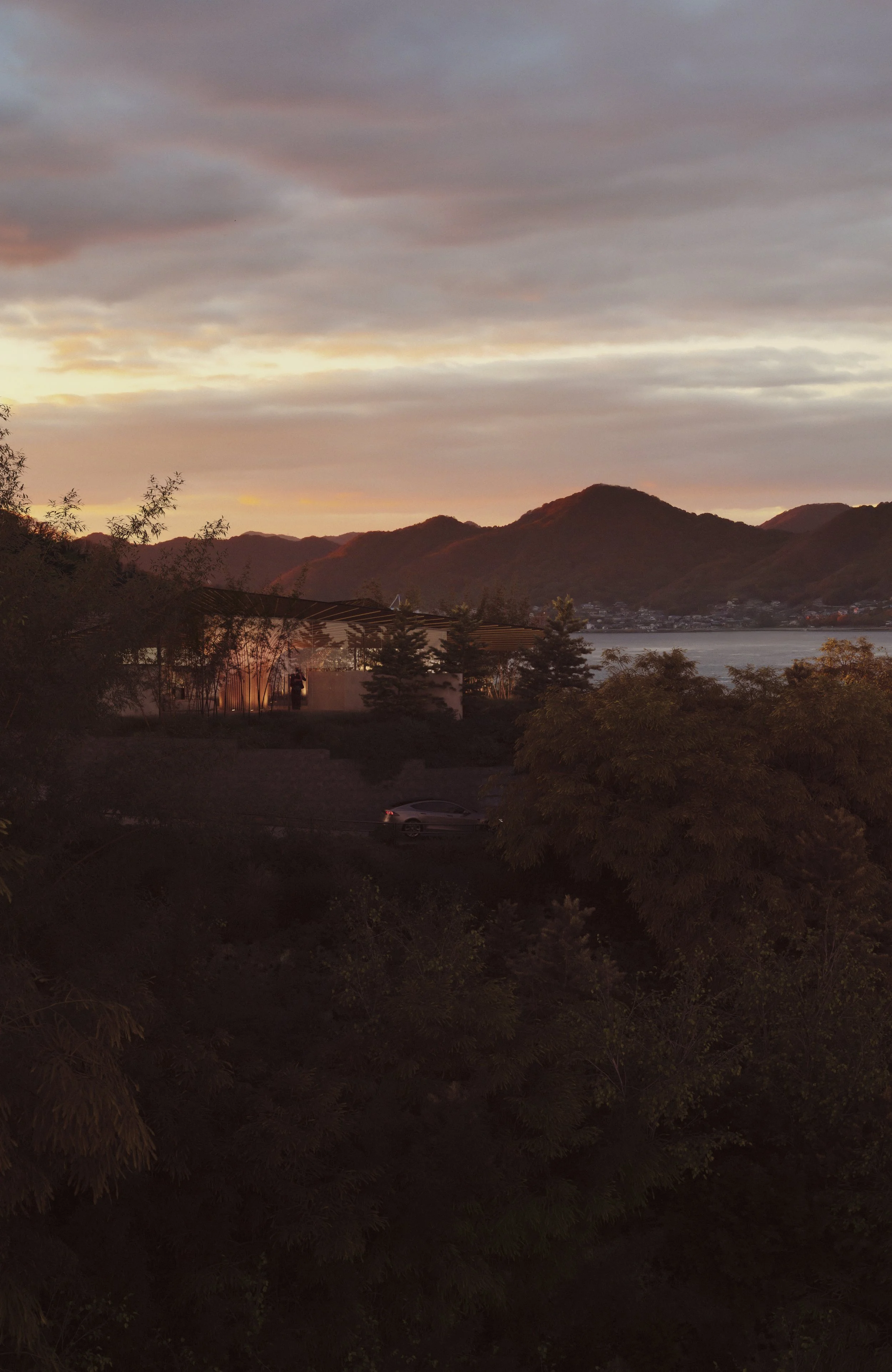 Scenic view of a sunset over mountains, with a lake in the background, trees in the foreground, and a building with lights on the left side.