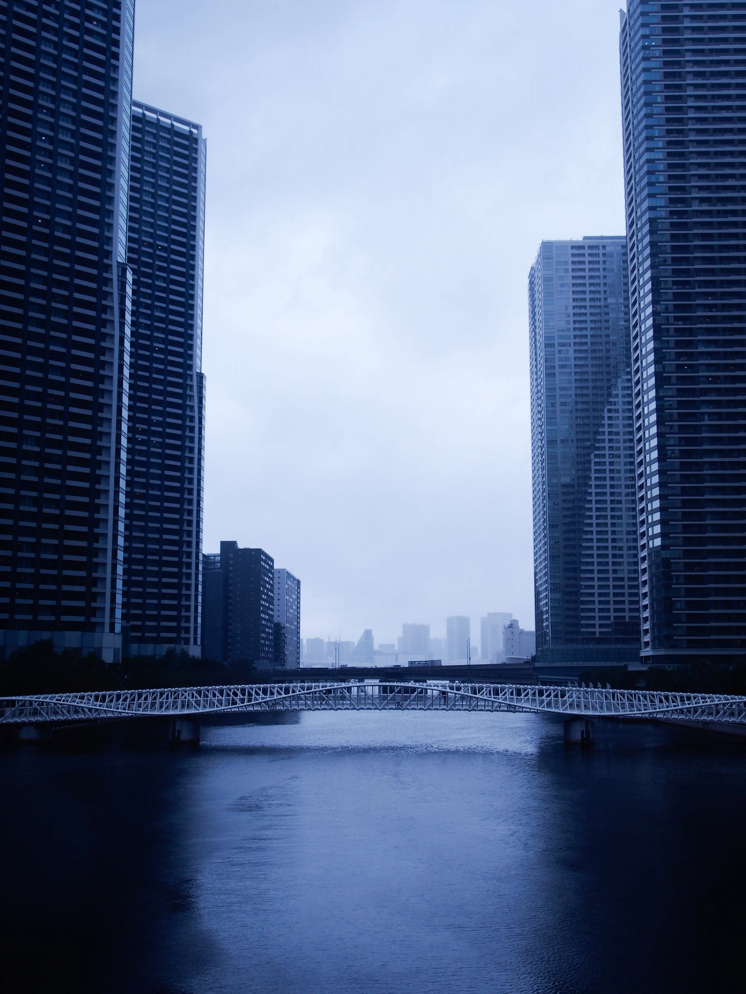 A cityscape with tall skyscrapers on either side of a river, a bridge crossing the river, and a cloudy sky overhead.