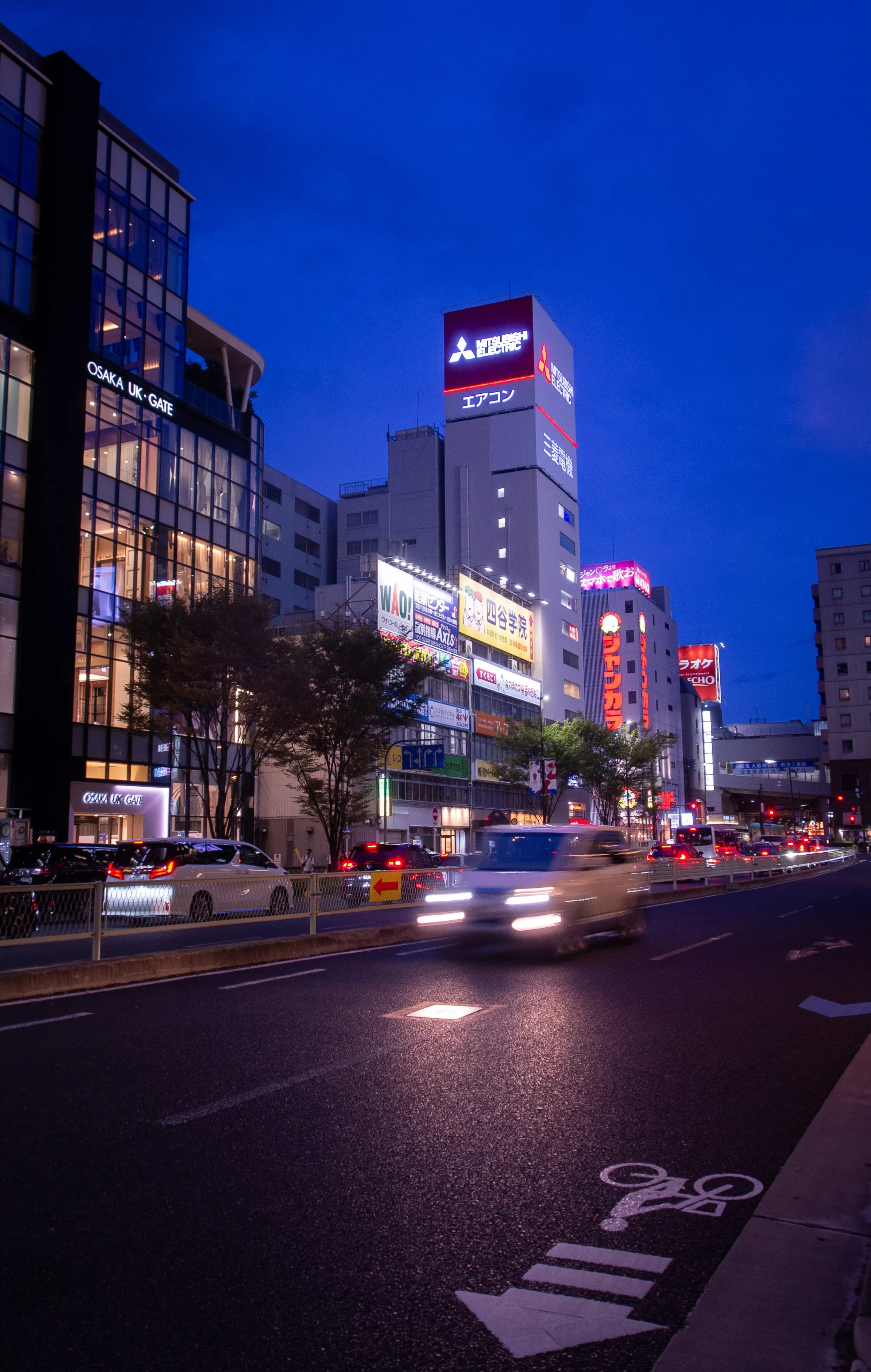 City street scene at dusk with illuminated buildings, advertisements, and moving vehicles on a wet road.