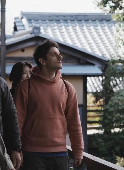 Young man in a brown hoodie standing outside near a traditional Japanese house with tiled roof, surrounded by trees.