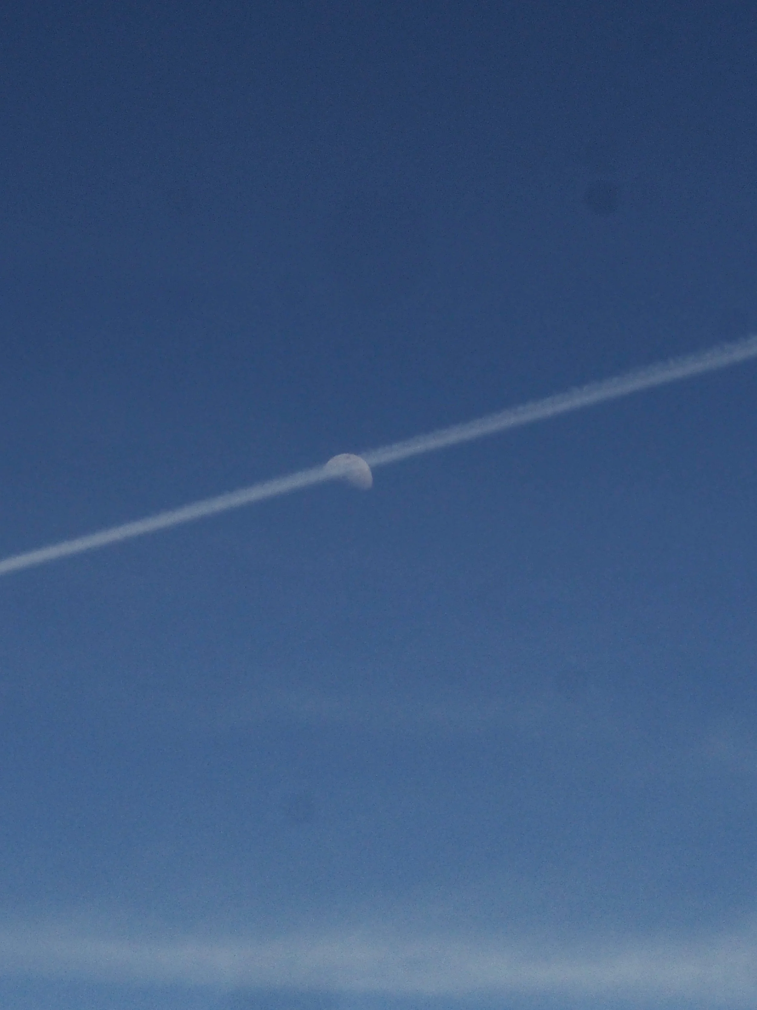 The moon partially visible in the blue sky with a contrail passing through it.