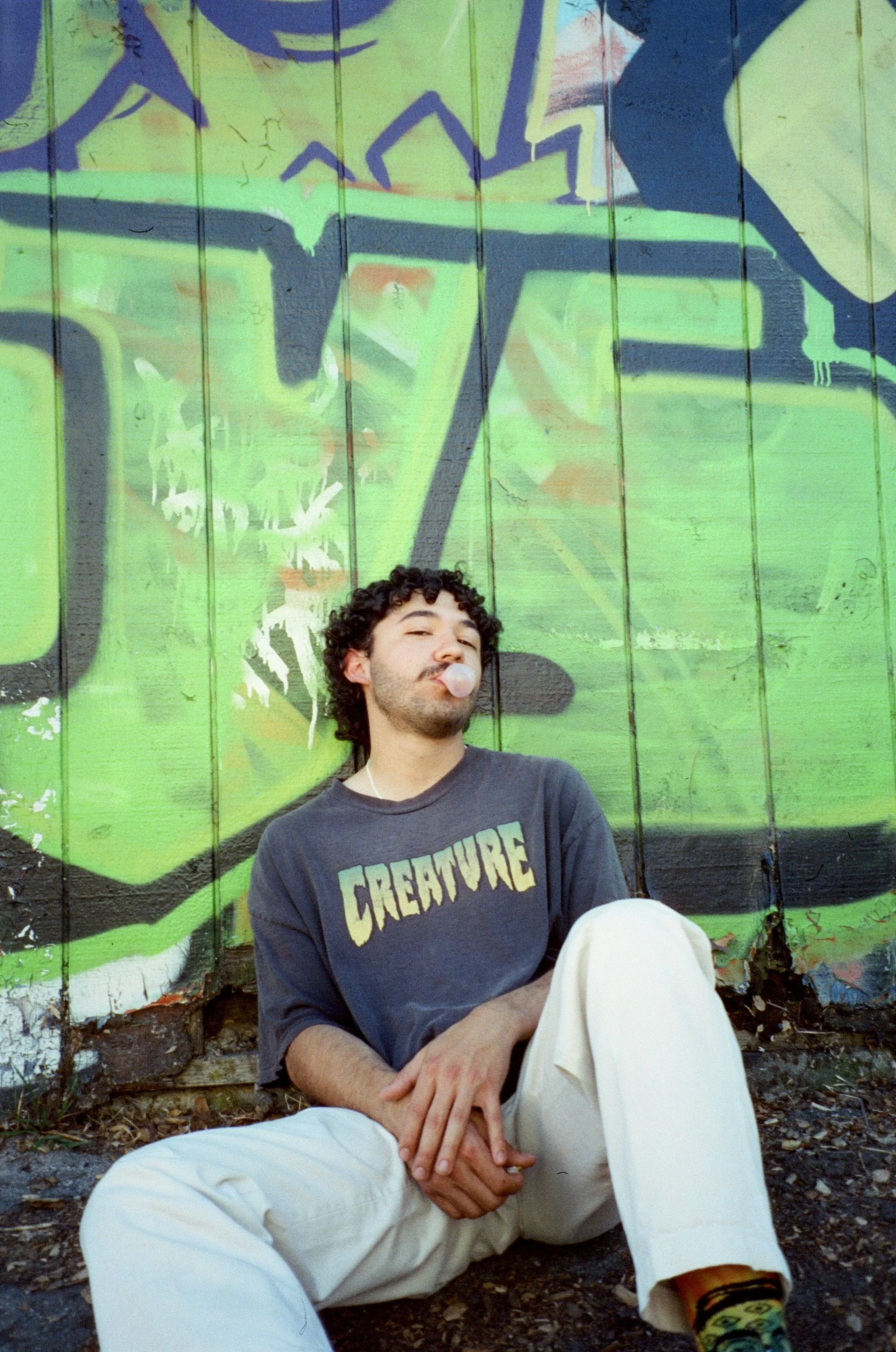 A young man with curly hair and a mustache is sitting against a green graffiti-covered wall. He is blowing a bubble gum bubble, wearing a dark T-shirt with the word 'CREATURE' on it, white pants, and colorful socks. The photo is taken outdoors on a gravel surface.