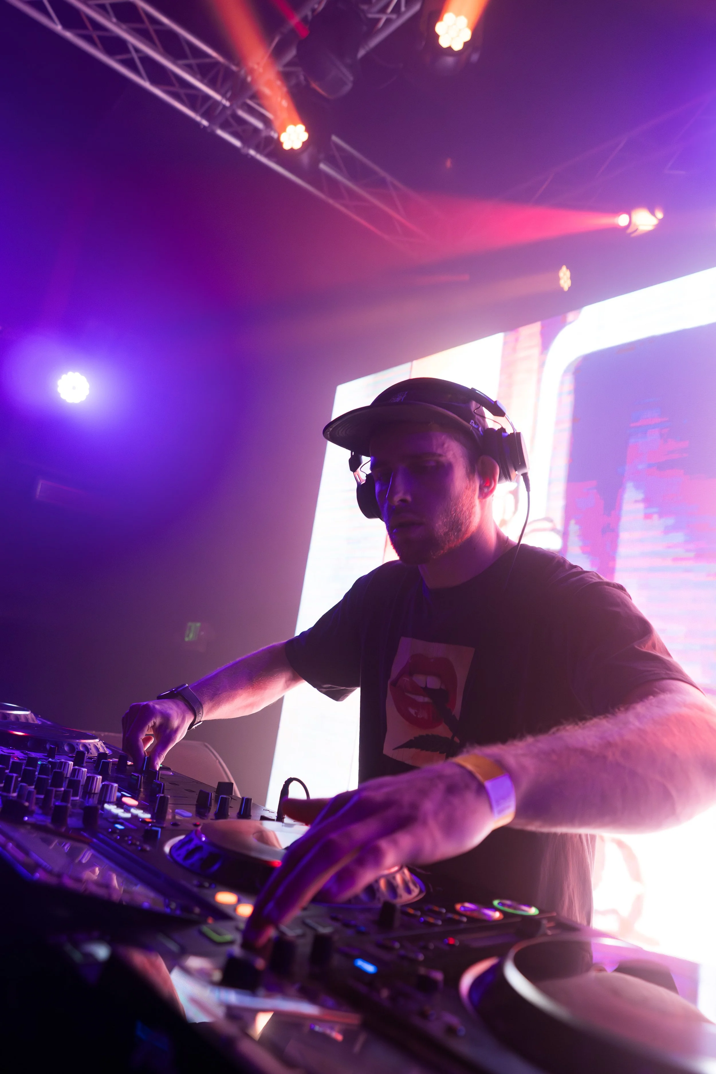 A DJ wearing a black cap and headphones is mixing music at a nightclub or concert, illuminated by colorful stage lights and a large LED screen in the background.