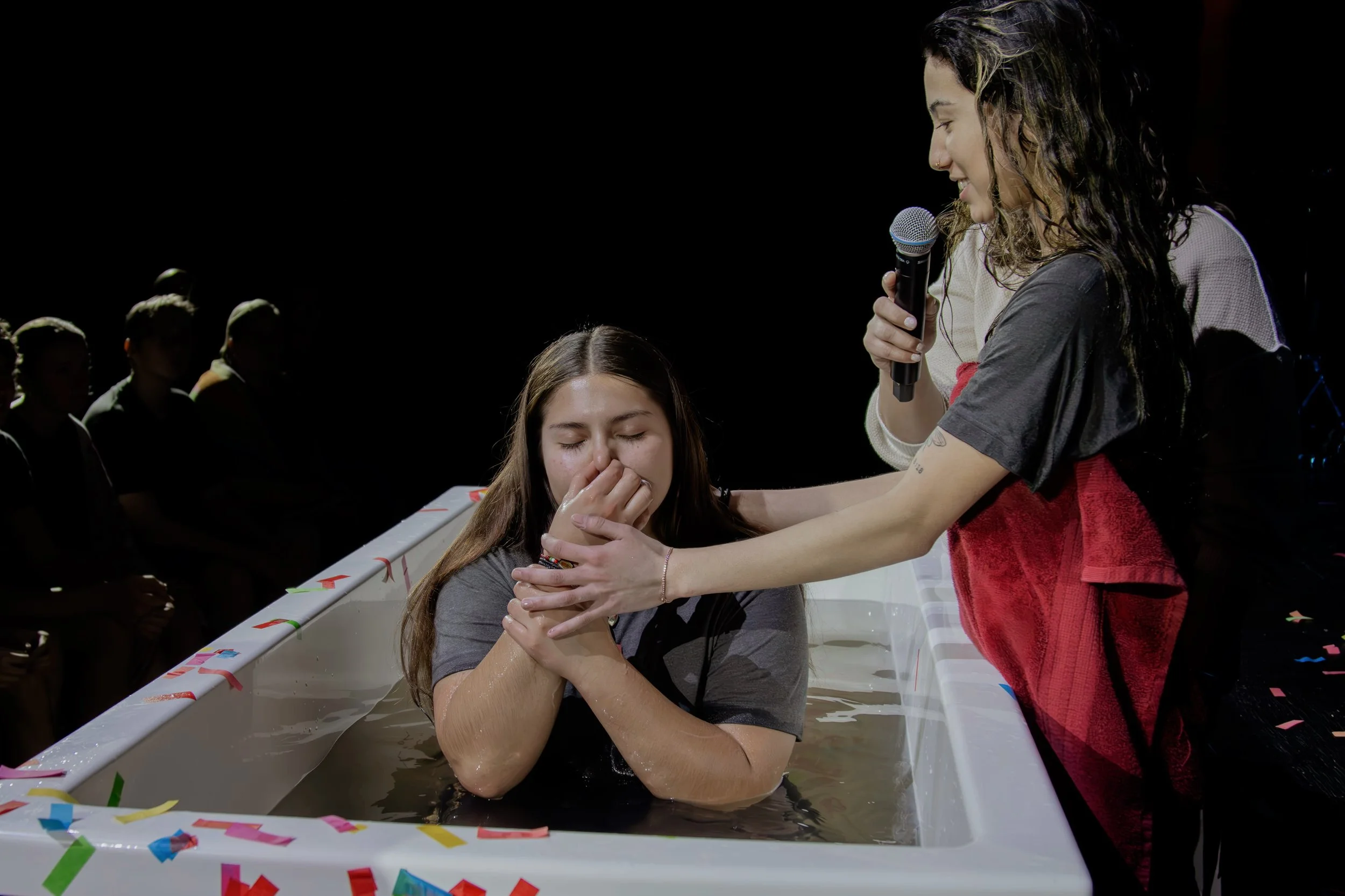 Young woman being baptized in a water tank while an assistant holds her hand and speaks into a microphone, with onlookers watching in the background.