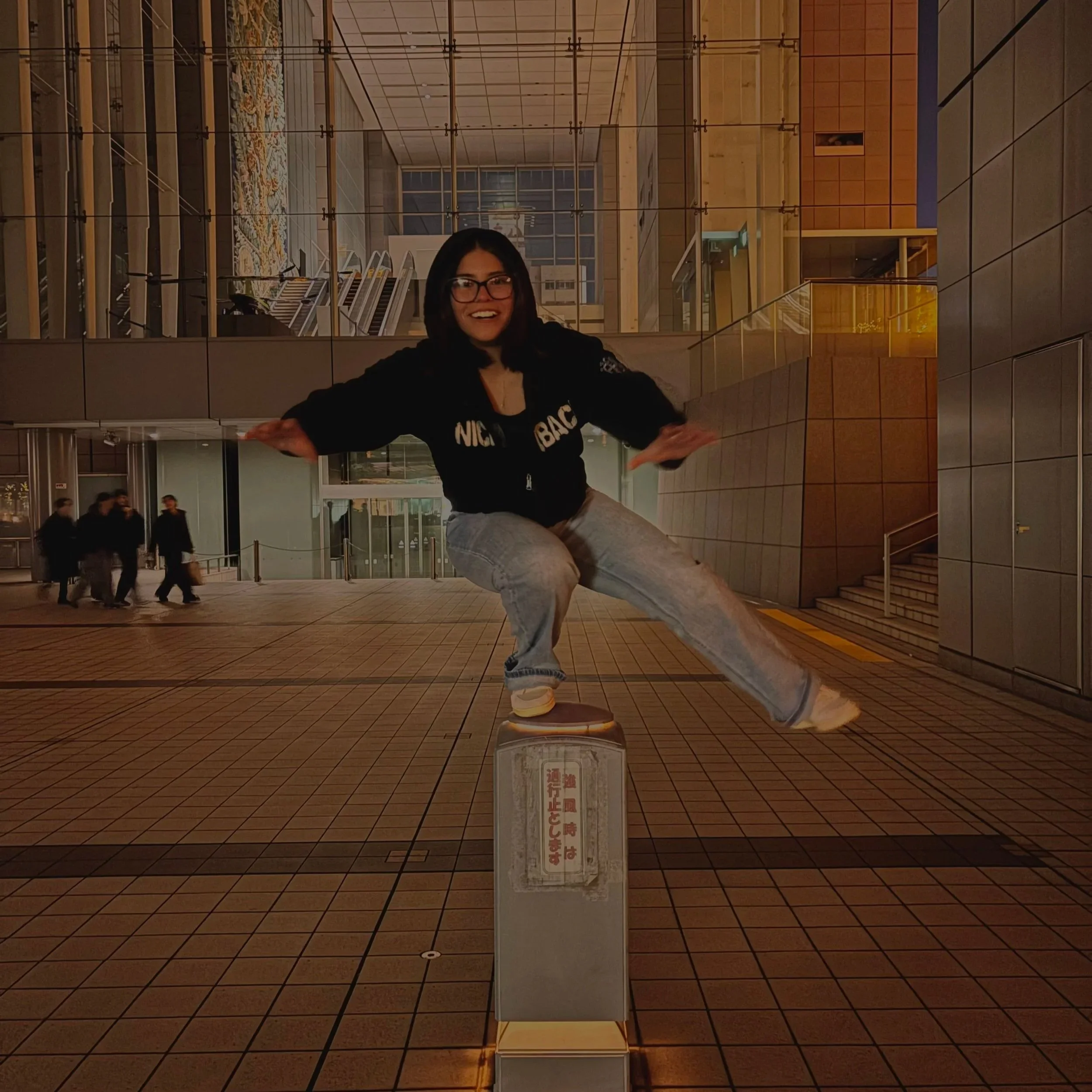 A woman with black hair and glasses balancing on a small pole in an urban area at night, with a glass building and people walking in the background.