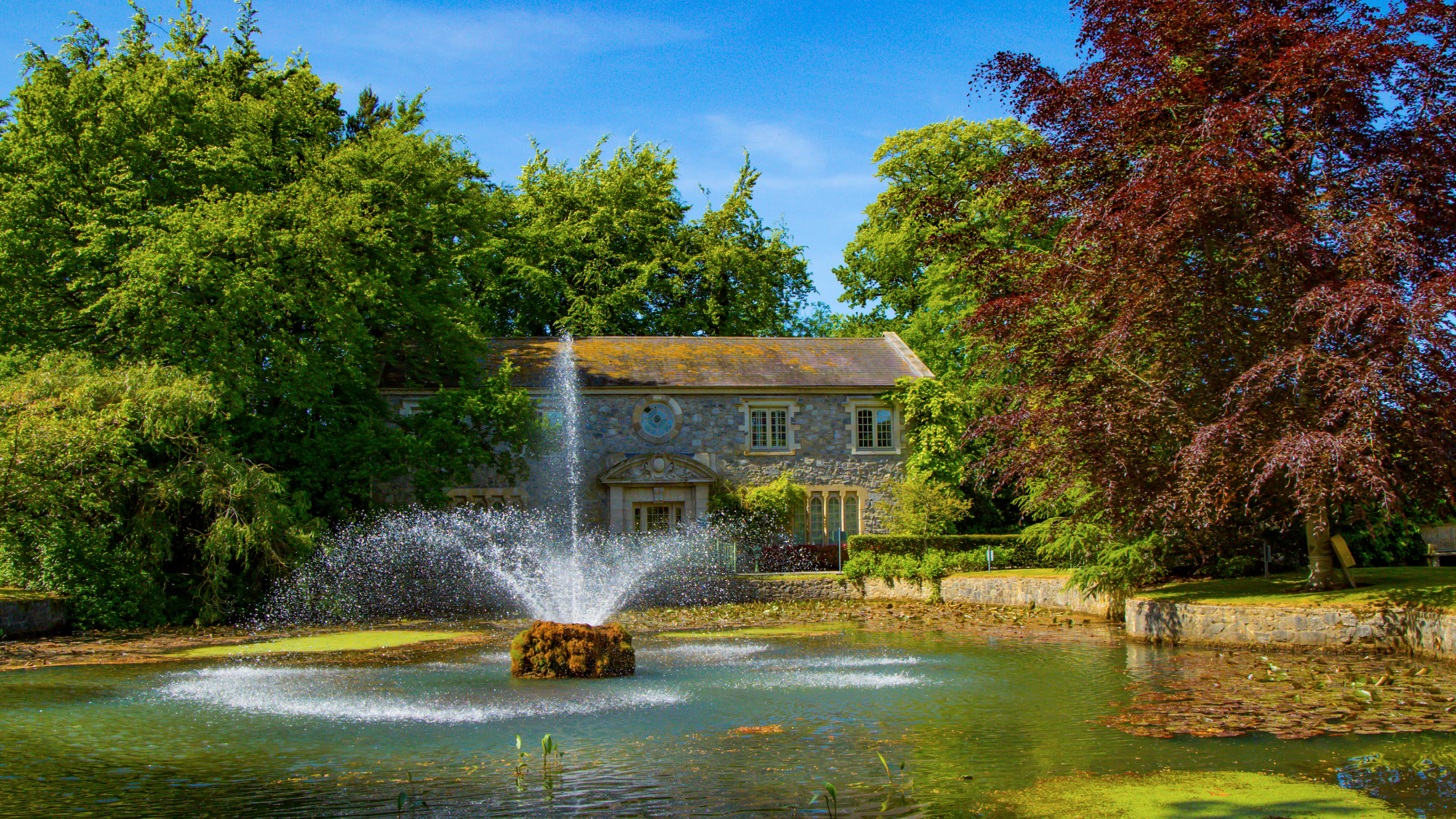 A stone house with a fountain in the pond in front, surrounded by lush green trees on a sunny day.