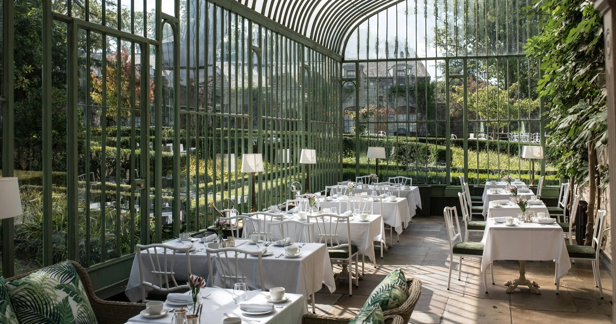 A bright, elegant dining area inside a glass-enclosed conservatory with white tablecloths, chairs, table settings, and potted plants, overlooking a lush garden outside.