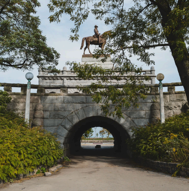 A stone arched tunnel with a statue of a person on a horse on top, surrounded by green trees under a cloudy sky.