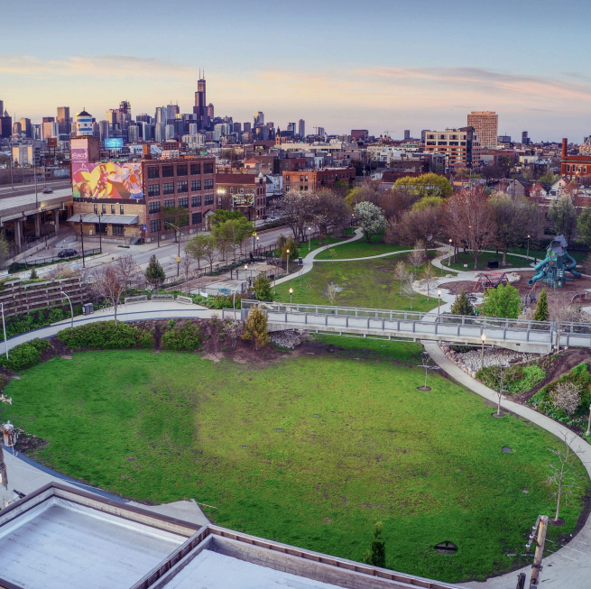 A city park with green grass, walking paths, trees, and playground equipment, with a skyline in the background under a partly cloudy sky.
