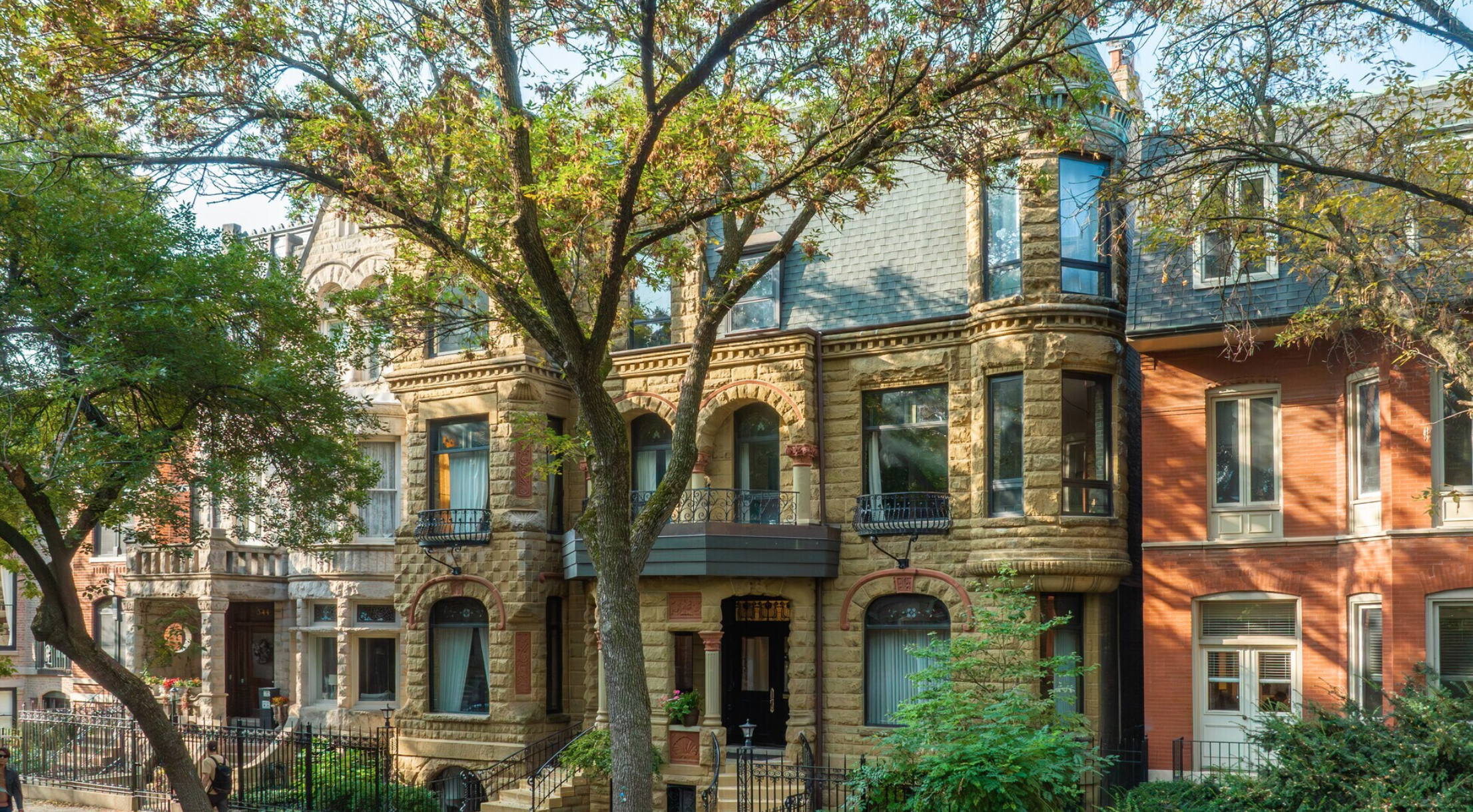 Row of historic residential buildings with Victorian-style architecture, stone facades, and large windows, partially obscured by trees with green leaves.