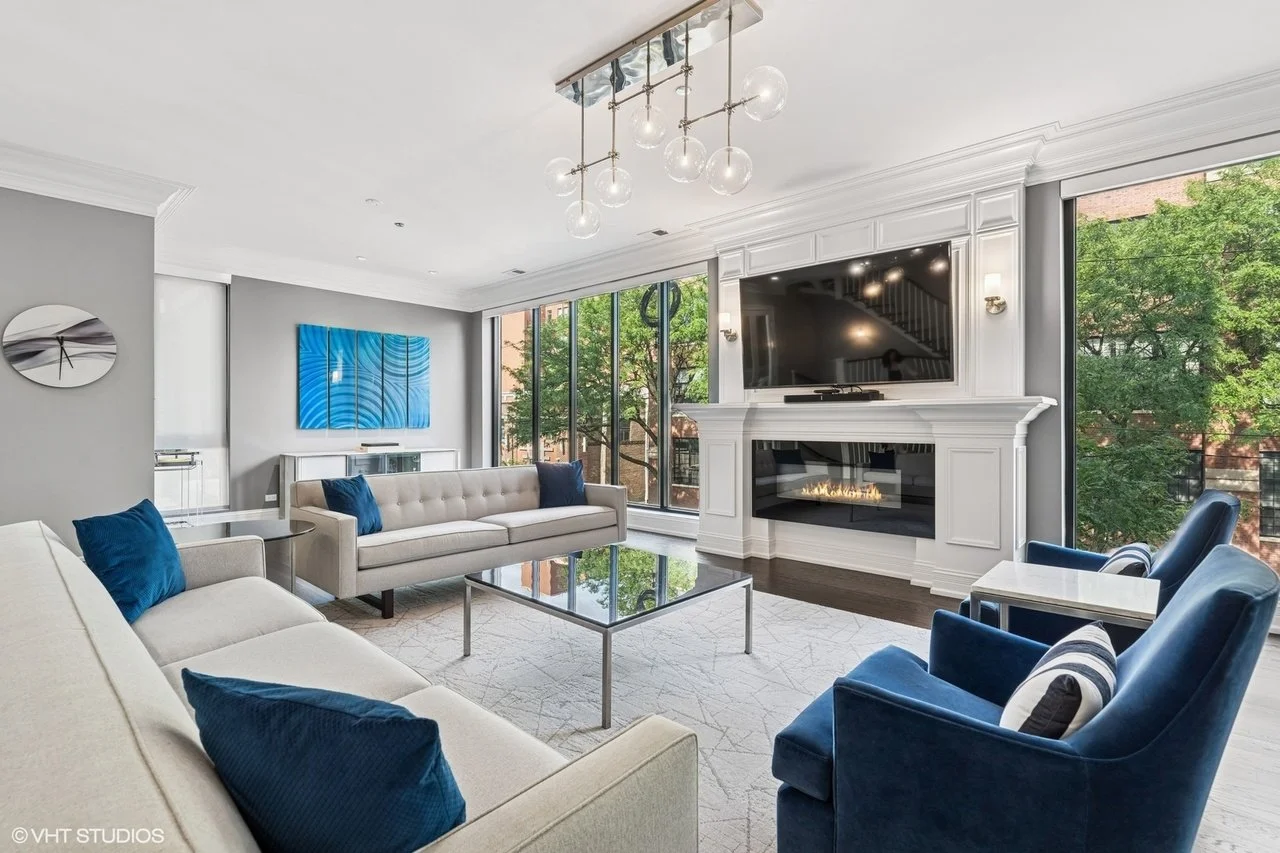 Modern living room with gray walls, large windows, white fireplace, flat-screen TV, beige and blue armchairs, a glass coffee table, and abstract art on the wall.