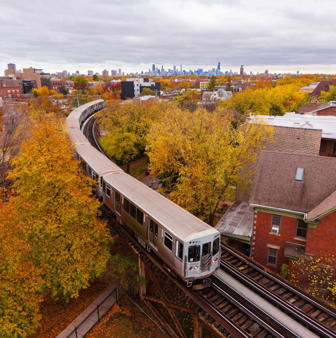 A train traveling on an elevated track through a city with autumn trees and a distant skyline.