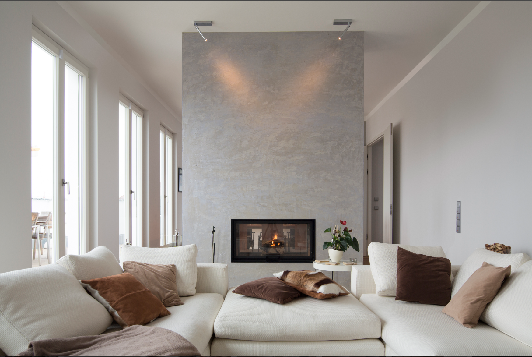 Living room with a fireplace on a textured wall, cream-colored sofa with brown and beige pillows, a small side table with a potted plant, and large windows letting in natural light.