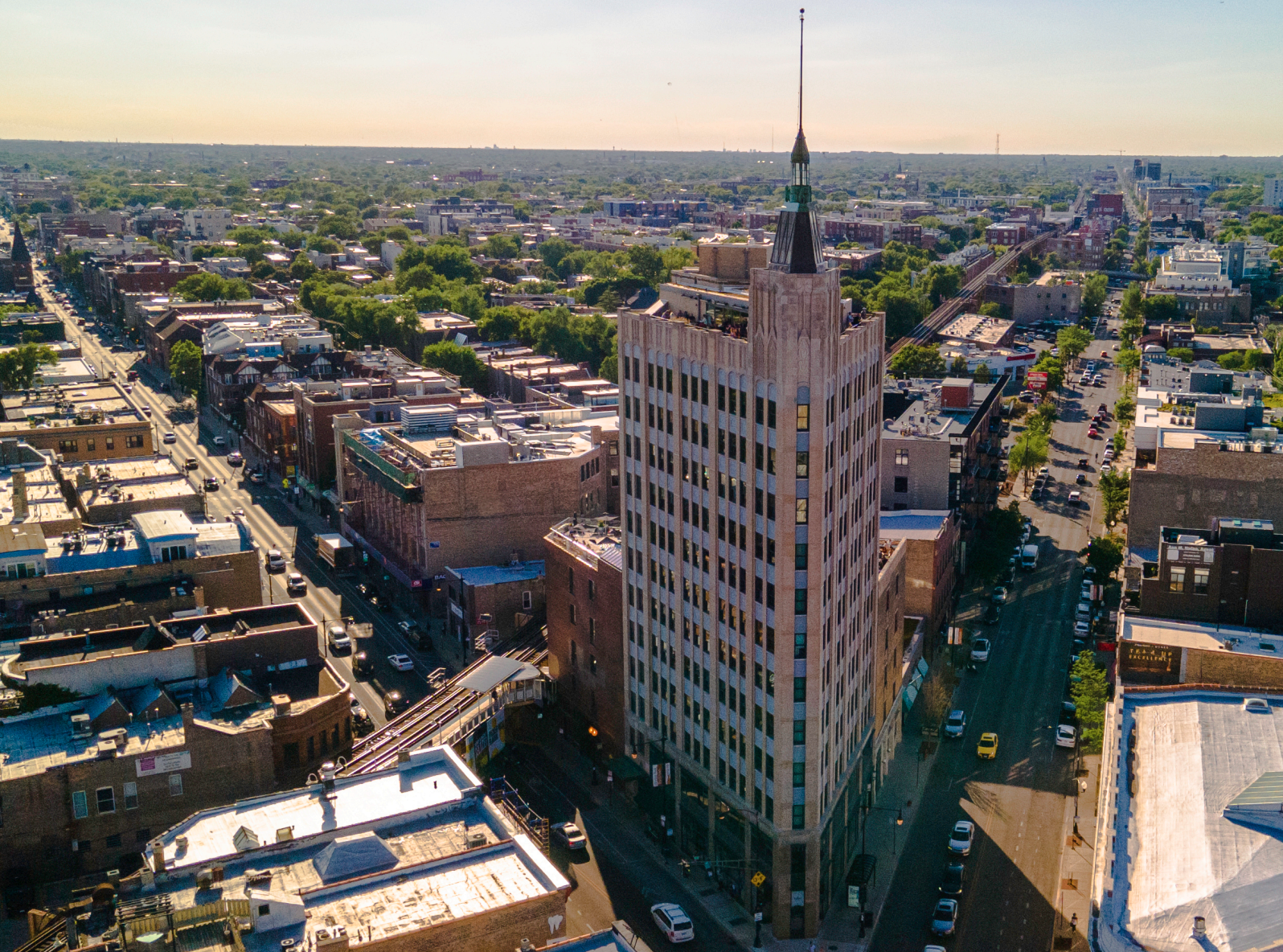 Aerial view of Bucktown featuring a tall, white building with a spire, The Robey, surrounded by other buildings, streets, and trees, under a clear sky during sunset.
