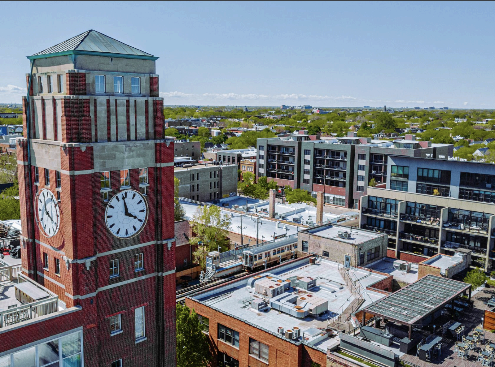 A cityscape featuring a tall red brick clock tower with white clock faces showing 2:55, surrounded by modern multi-story buildings with rooftop HVAC units and a rooftop terrace, under a clear blue sky.