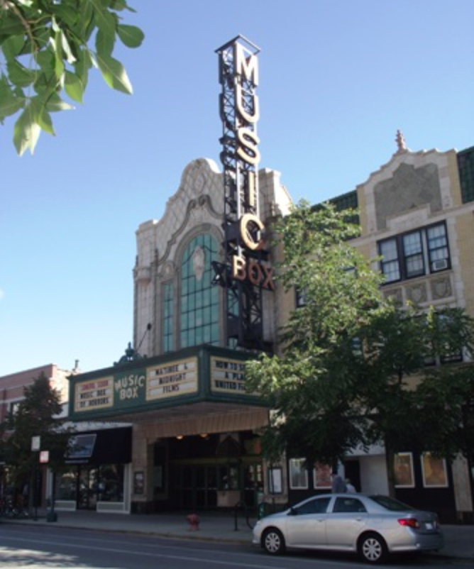 Sign for the Music Box theater on a building with a marquee, storefront, and street view with parked cars and trees.