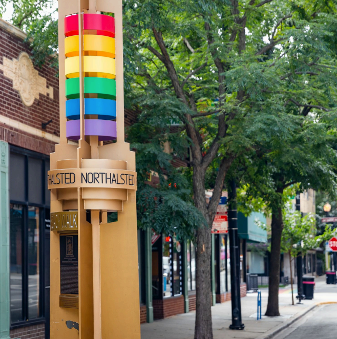 A colorful streetlight with rainbow-colored rings on a beige pole, with a tree and storefronts in the background.