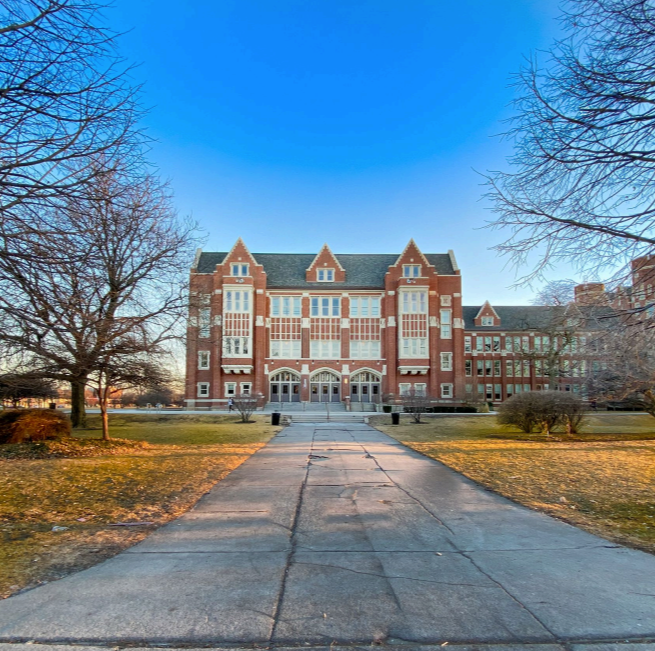 A large brick university building with gothic architectural features, surrounded by leafless trees and a paved walkway leading up to the entrance, under a clear blue sky.