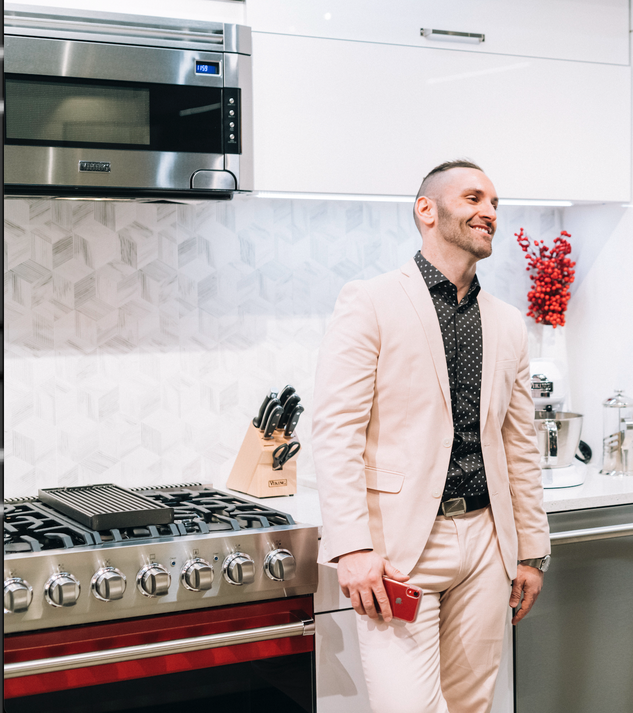 A man dressed in a light-colored suit holding a red phone, standing in a modern kitchen.
