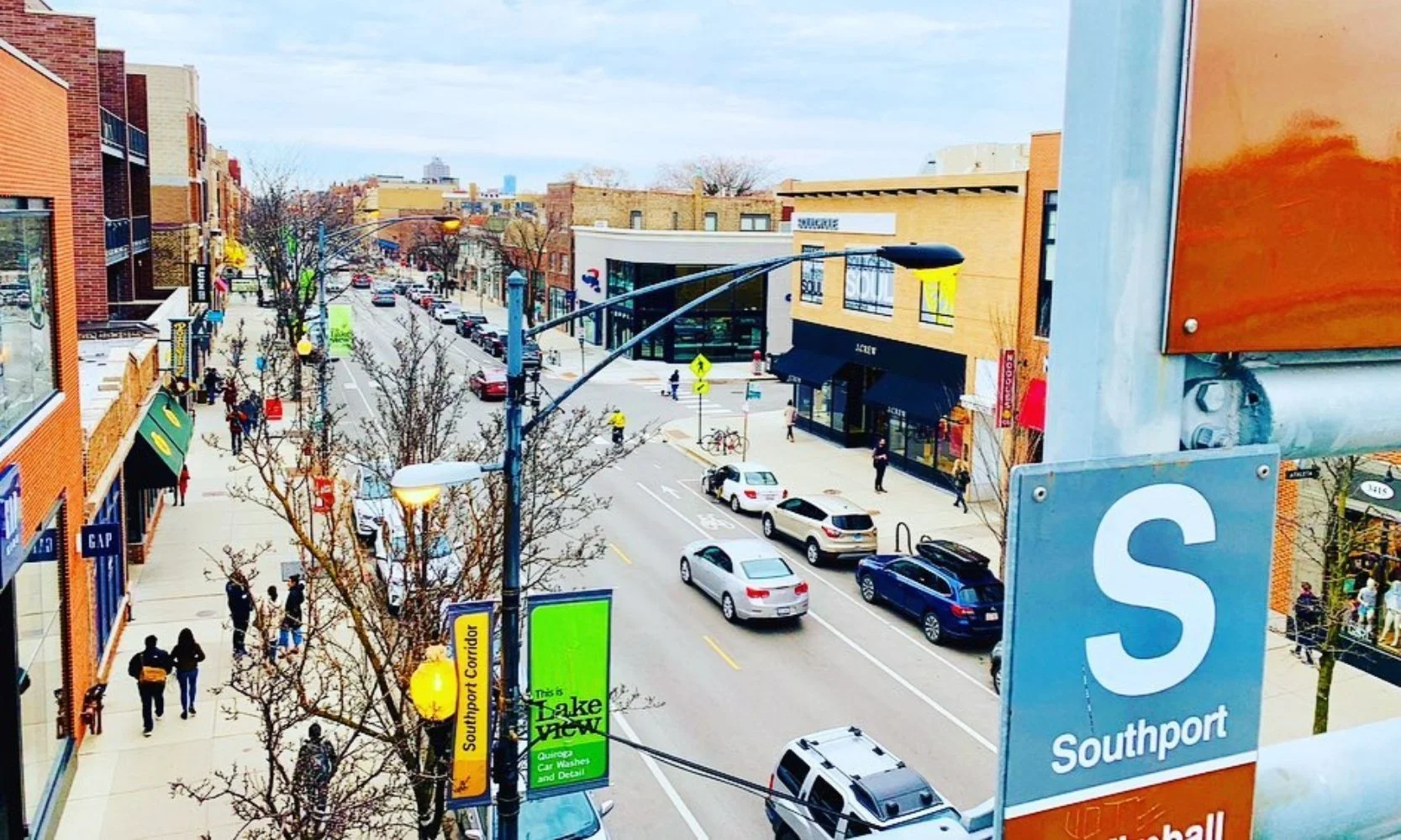 View of a busy downtown street with cars parked along the curb, storefronts on both sides, leafless trees, and pedestrians walking on the sidewalk. There are street lamps and signage, including a large blue sign that reads 'Southport' and a green sign for a car wash.