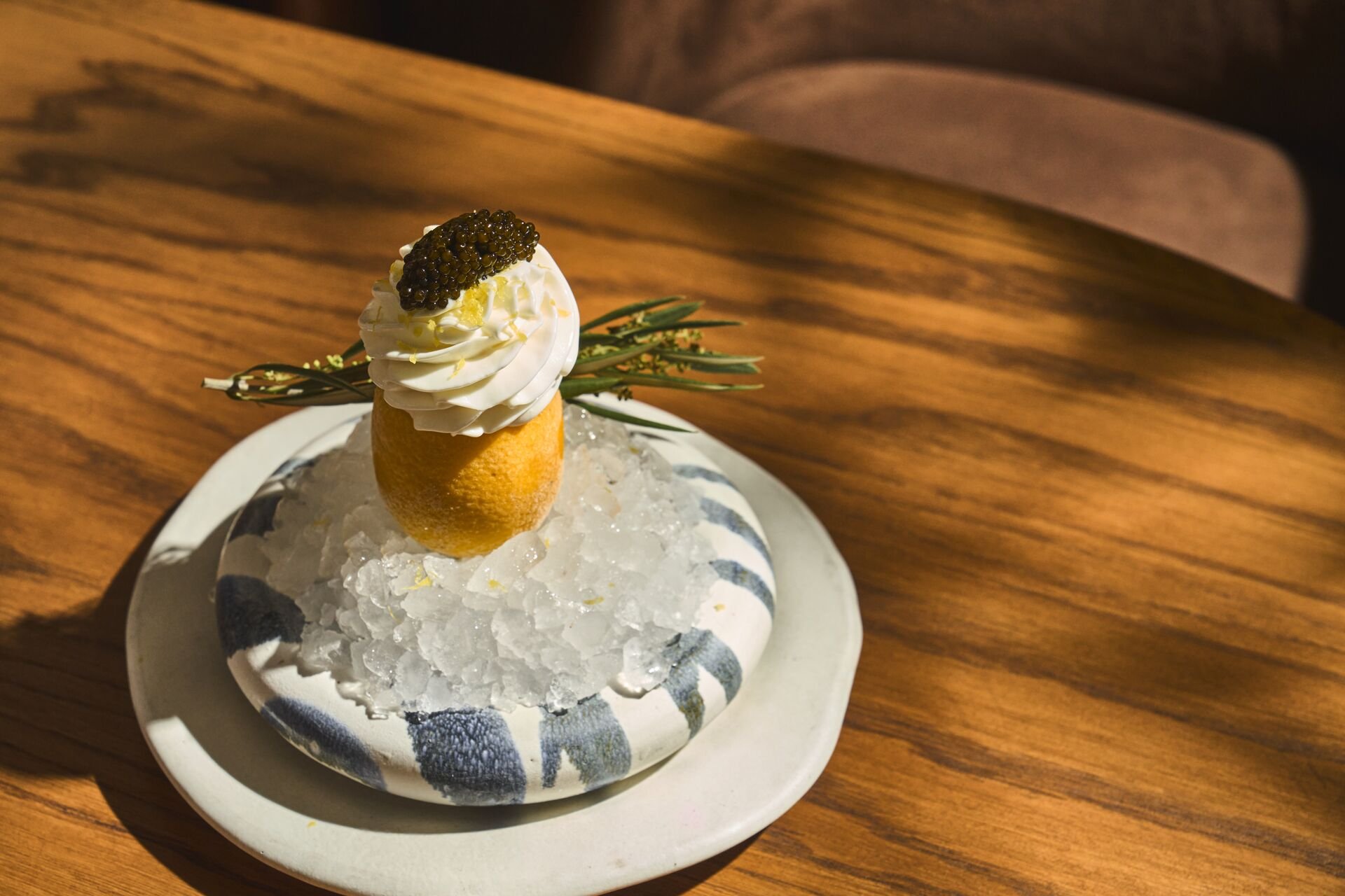 A dessert resembling a small cake or pastry topped with whipped cream and a spoonful of black caviar, garnished with a sprig of greenery, served on a white plate with ice and placed on a wooden table.