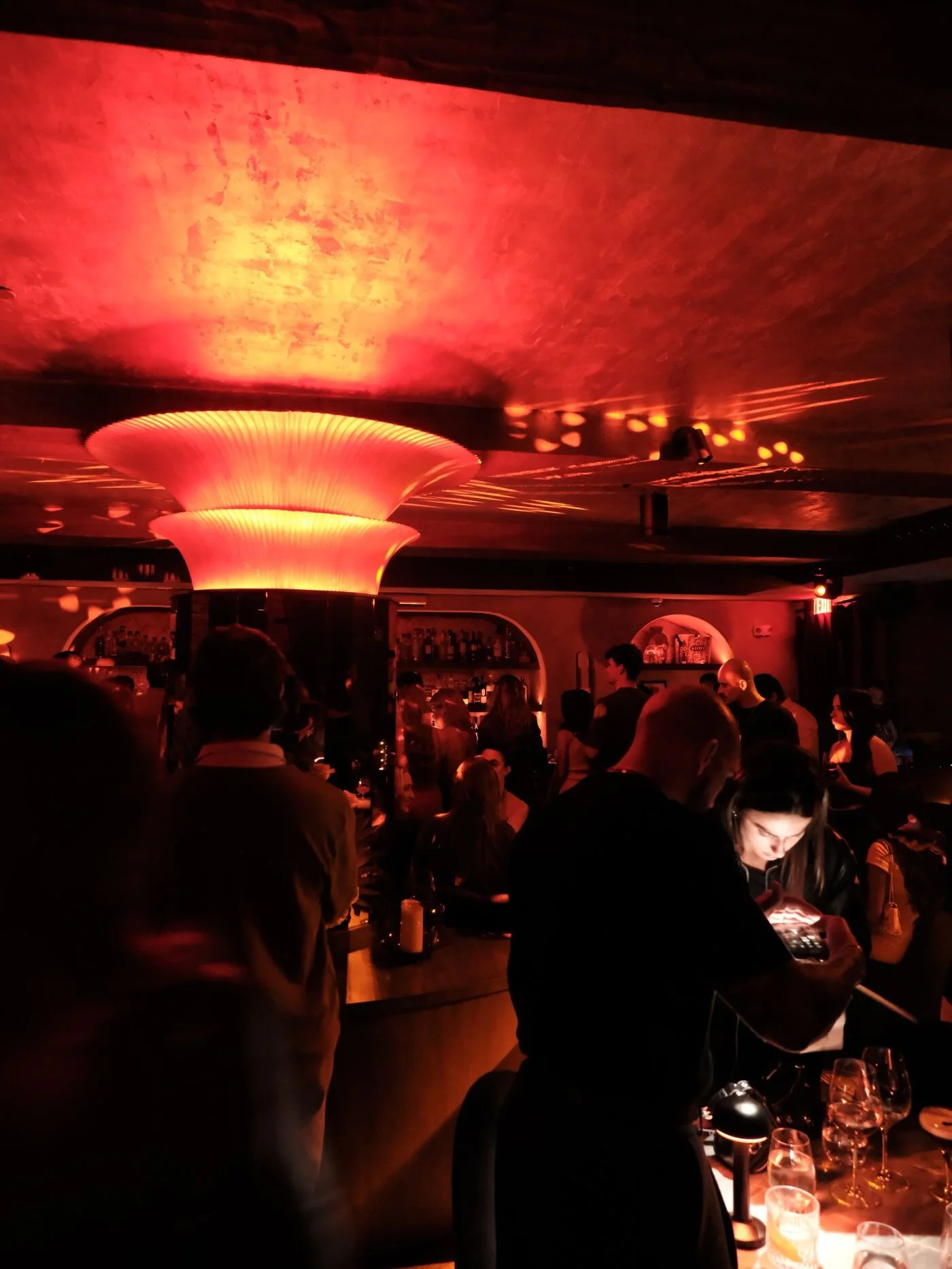 Dimly lit bar or restaurant with a large glowing red and orange light fixture in the ceiling. People are standing and sitting at tables, some using their phones. There are various glasses and bottles on the tables, and the background shows a bar area with shelves of bottles and glasses.