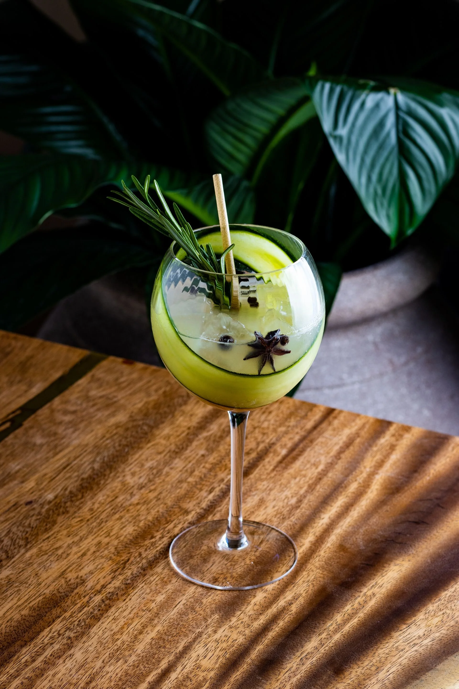 A cocktail glass with a lime wheel, star anise, rosemary, and a stir stick, placed on a wooden surface with green tropical plants in the background.