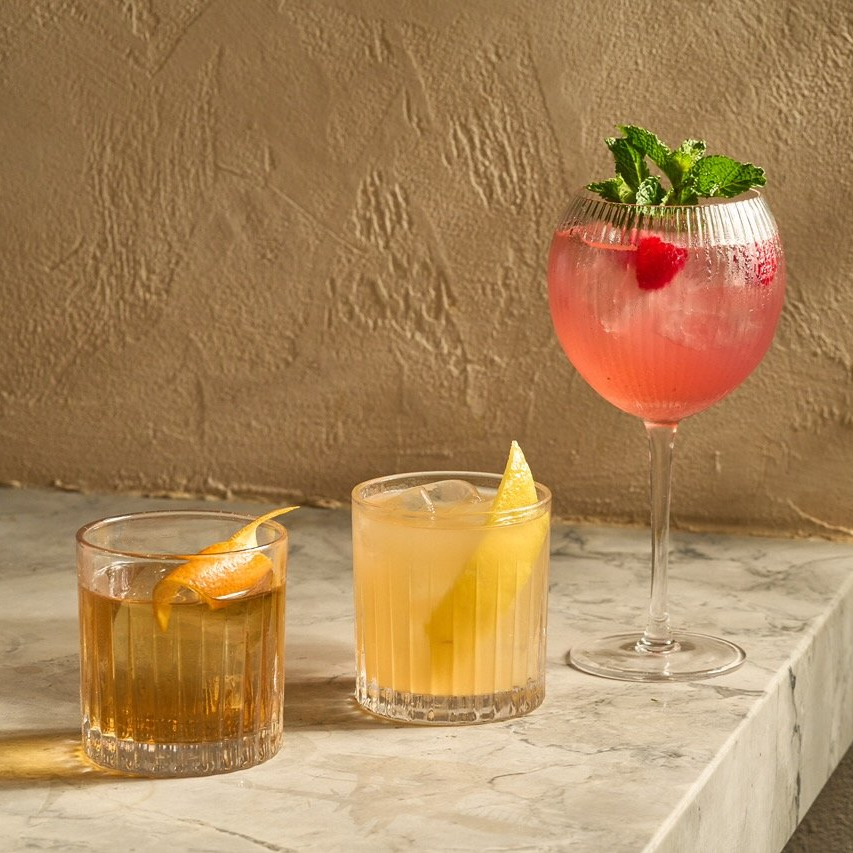 Three cocktails on a marble surface against a textured beige wall. The first is a short drink with an orange peel garnish, the second is a yellow lemonade with lemon slices, and the third is a pink cocktail with raspberries and mint leaves in a tall glass.