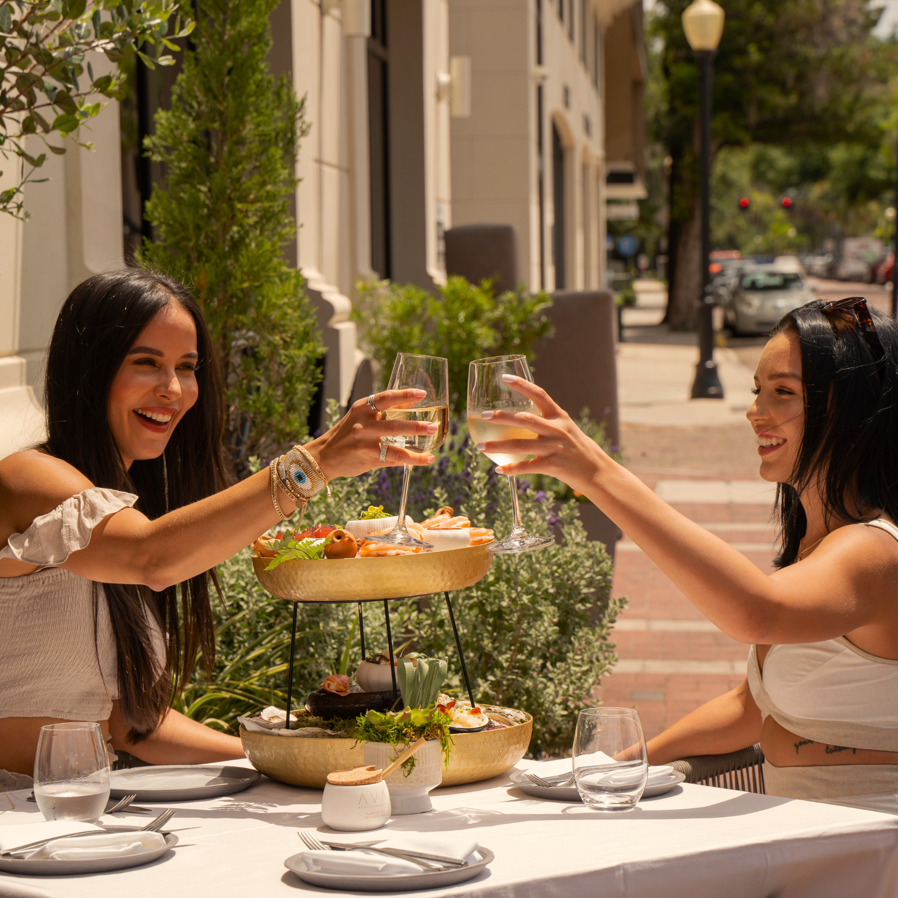 Two women clinking glasses of white wine at an outdoor restaurant table with a multi-tiered serving tray of appetizers, surrounded by greenery and urban street setting.