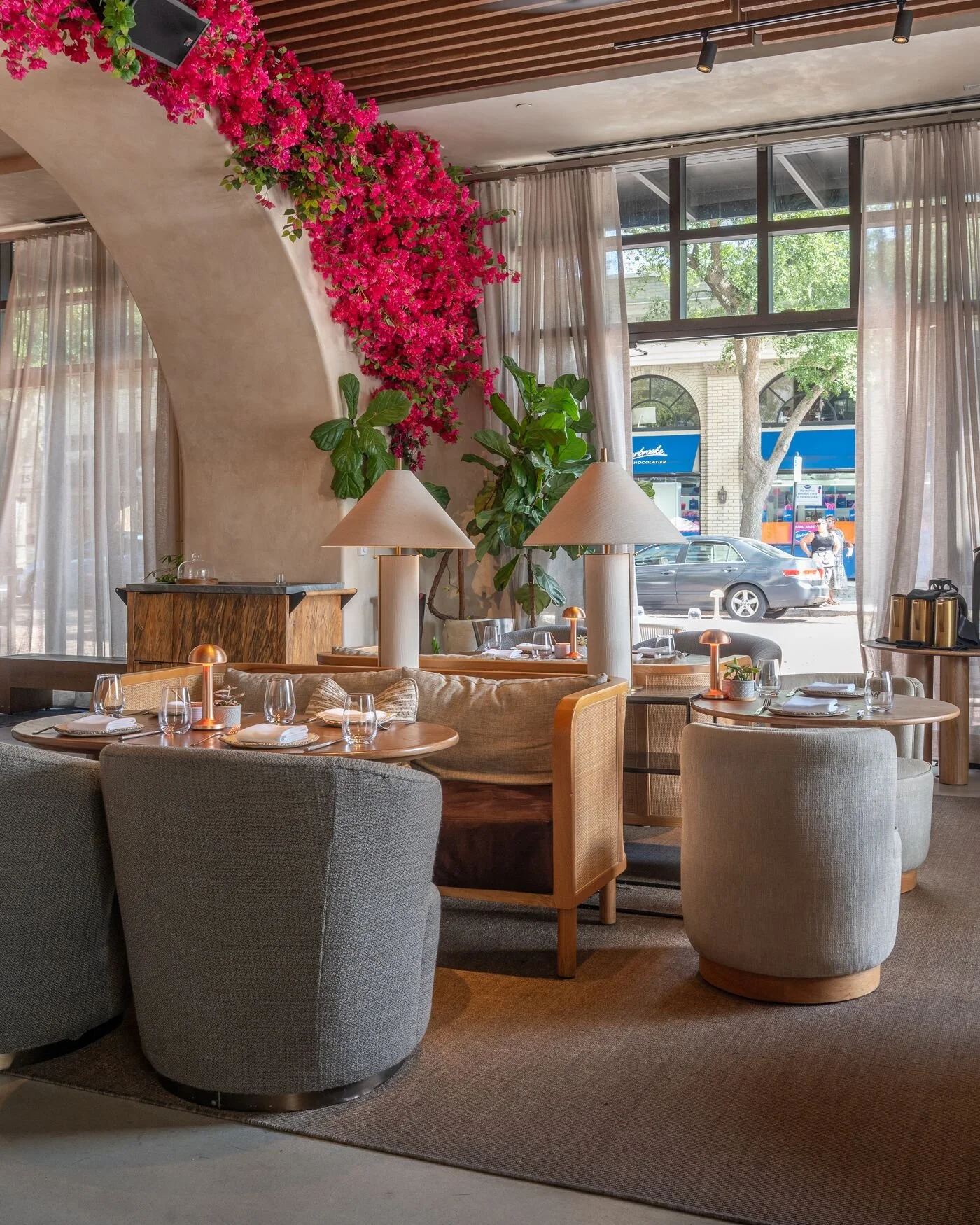 Dining area in a restaurant with a curved plaster wall decorated with pink bougainvillaea, large green indoor plants, beige and gray upholstered chairs, round tables set with glassware and napkins, cream-colored curtains, and large windows showing a 